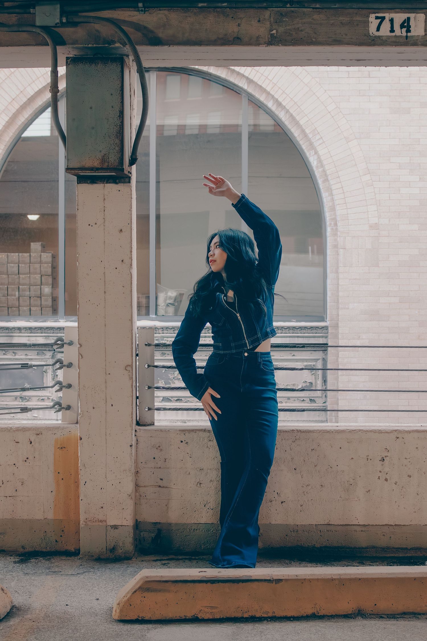 Woman in navy blue flare jeans and jacket poses with raised arm beside arched window in parking structure.