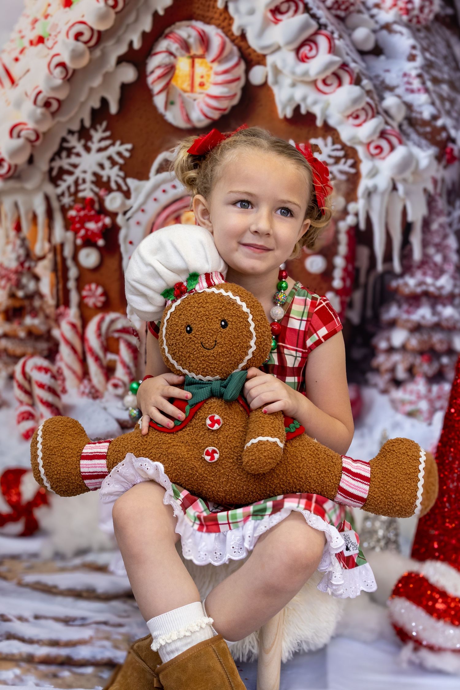 Young girl in plaid dress hugging a gingerbread man stuffed toy in front of a festive candy Christmas house.