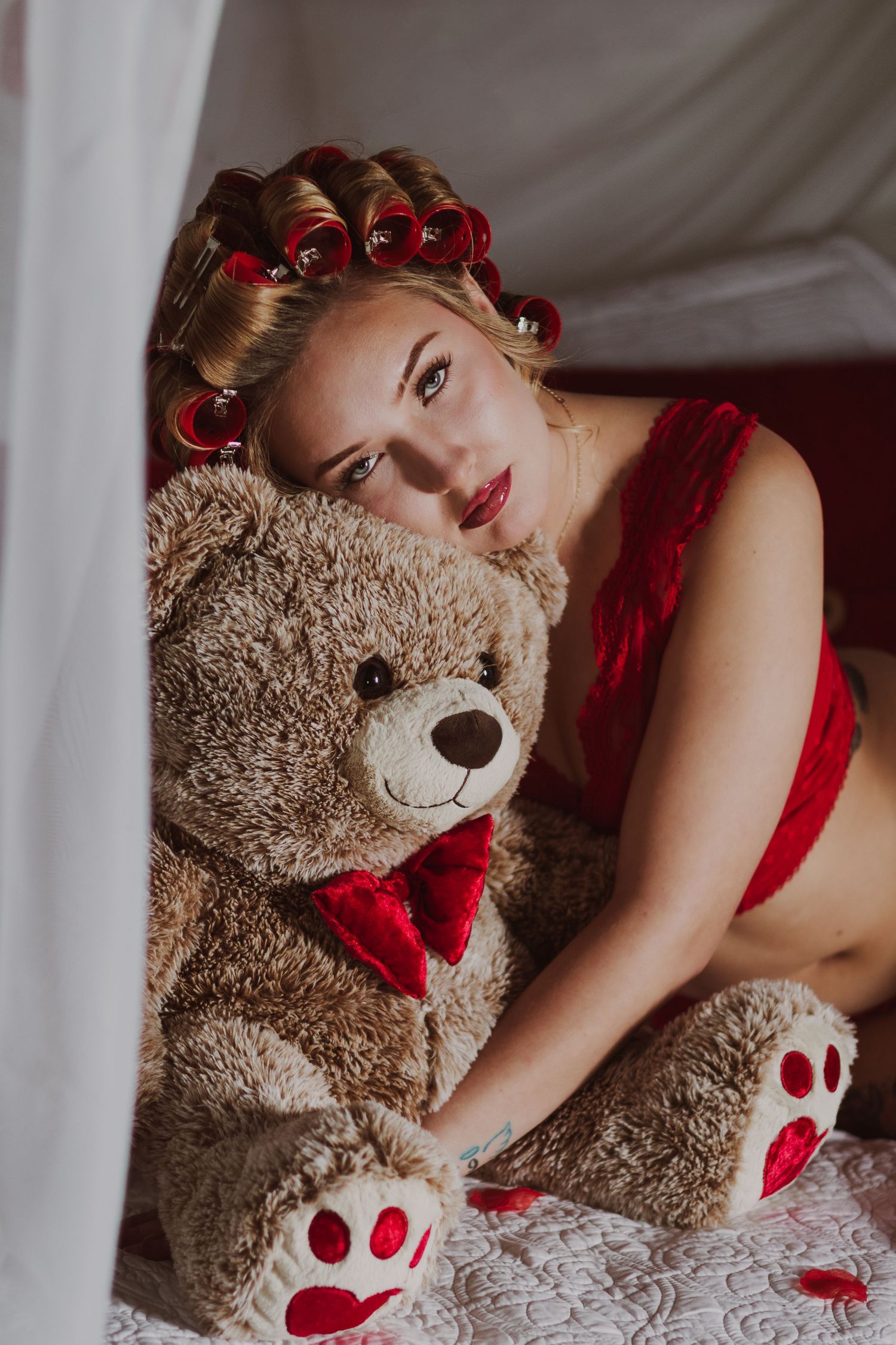 Blonde woman in red lingerie with hair curlers cuddling a large teddy bear on a white bed.
