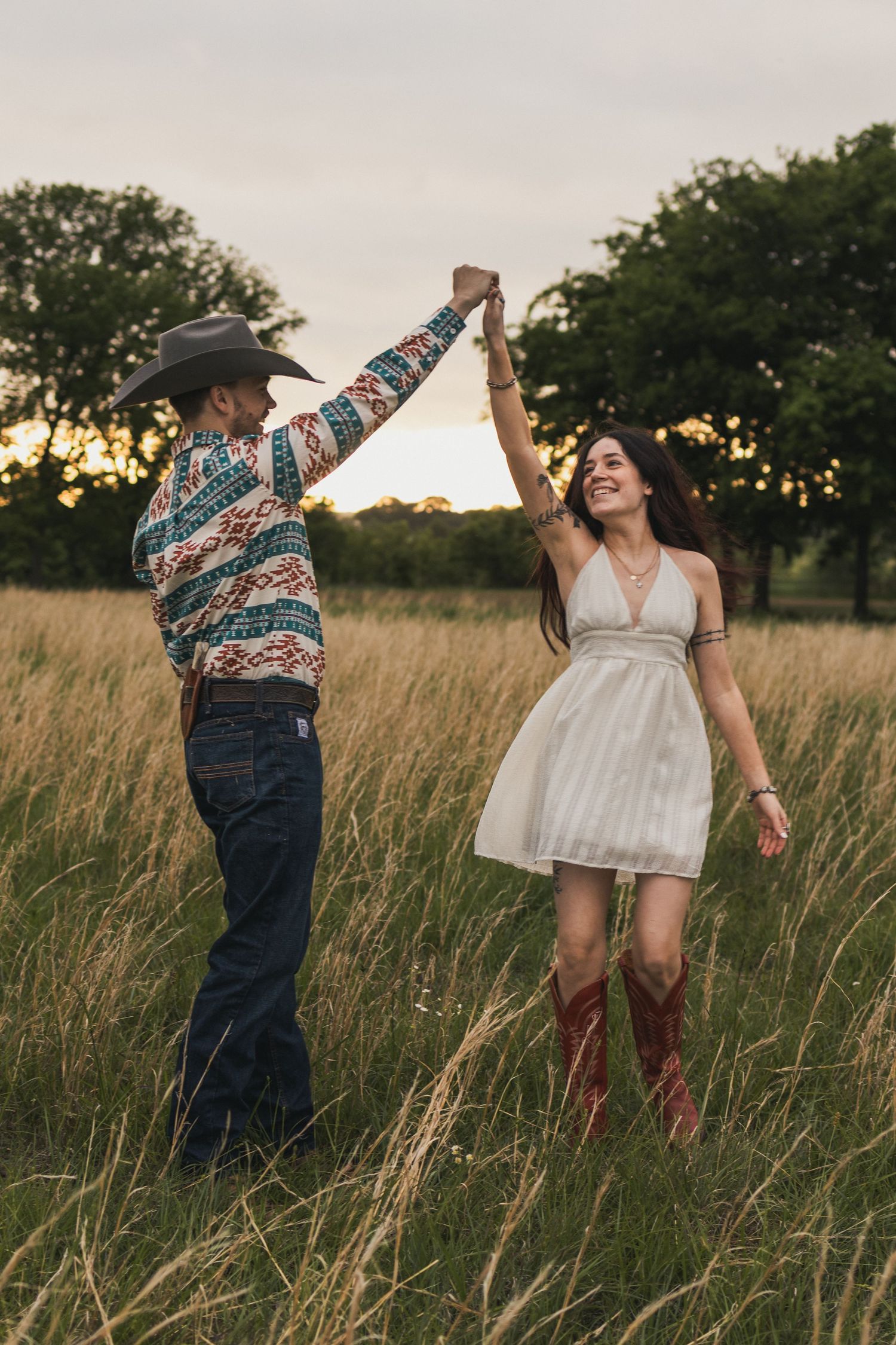 Couple dancing at sunset in tall grass field, man in cowboy hat and colorful shirt spinning woman in white dress and red boots.