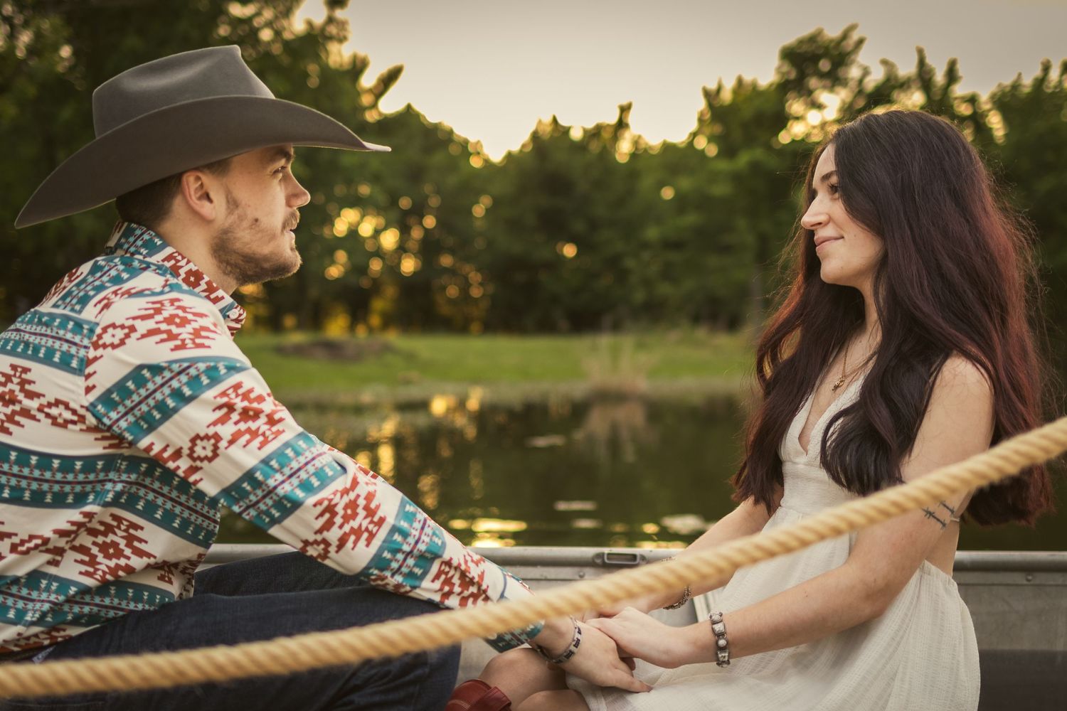 A couple in a rowboat holds hands near a sunlit pond, man in cowboy hat and patterned jacket, woman in white dress.