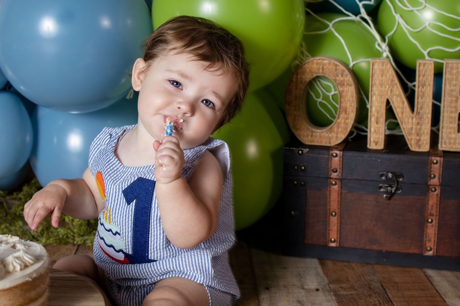 Happy baby boy in number 1 romper sits among blue and green balloons next to ONE wooden sign during birthday photoshoot.