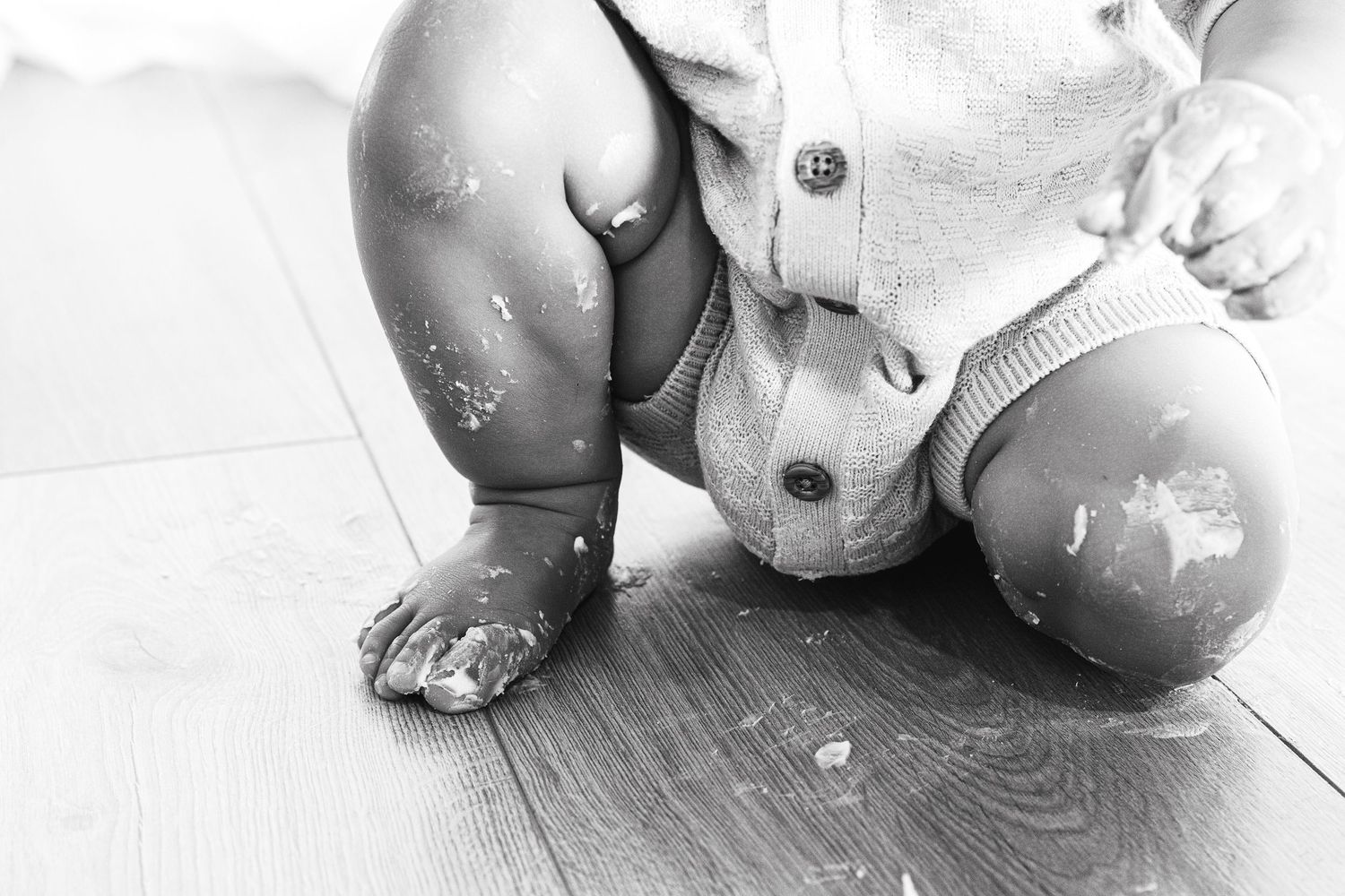 Black and white photo of a baby crawling on hardwood floor with messy cake on legs and onesie.