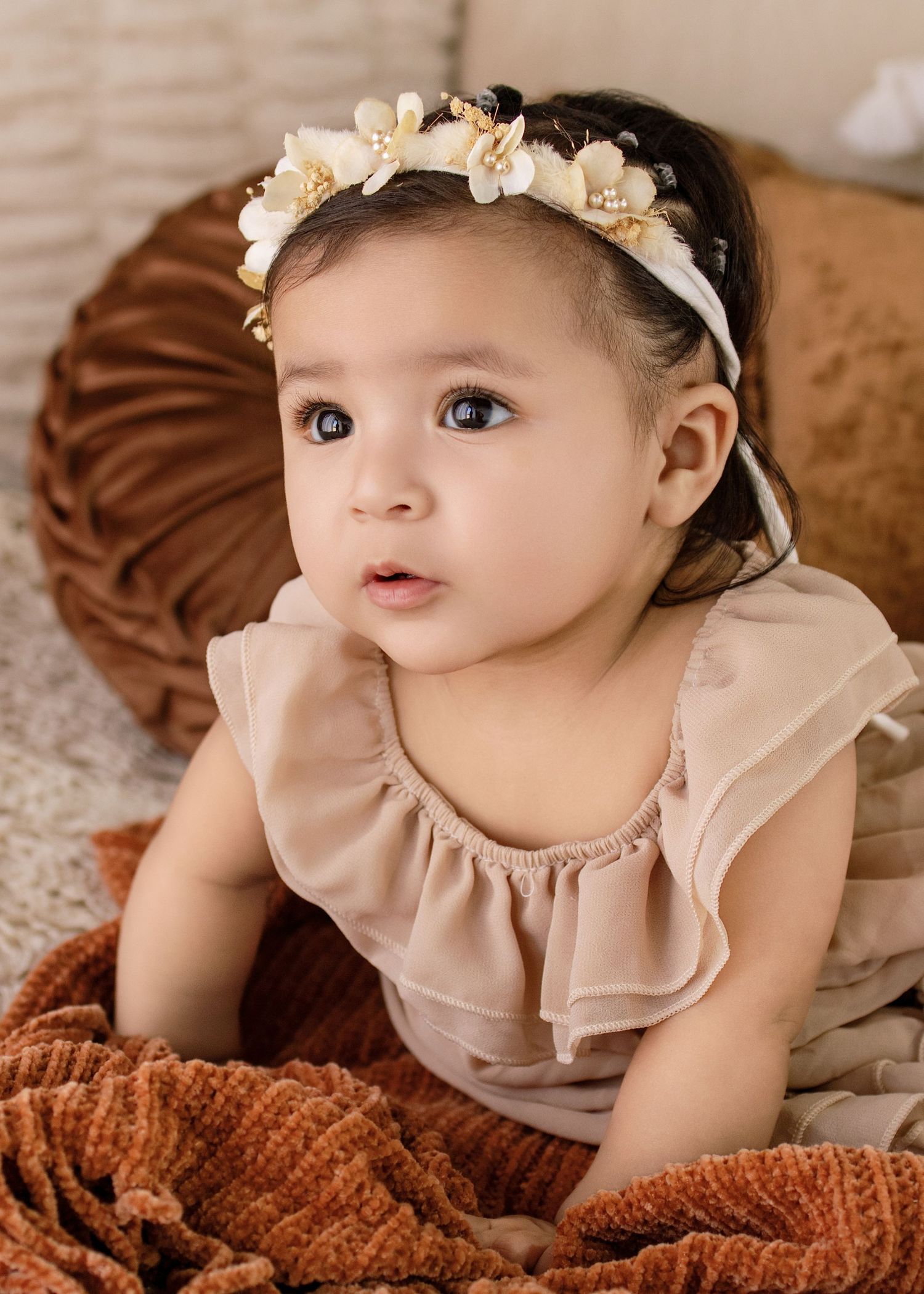 Adorable baby girl in a beige ruffle outfit and floral pearl headband, sitting on a warm brown velvet cushion.