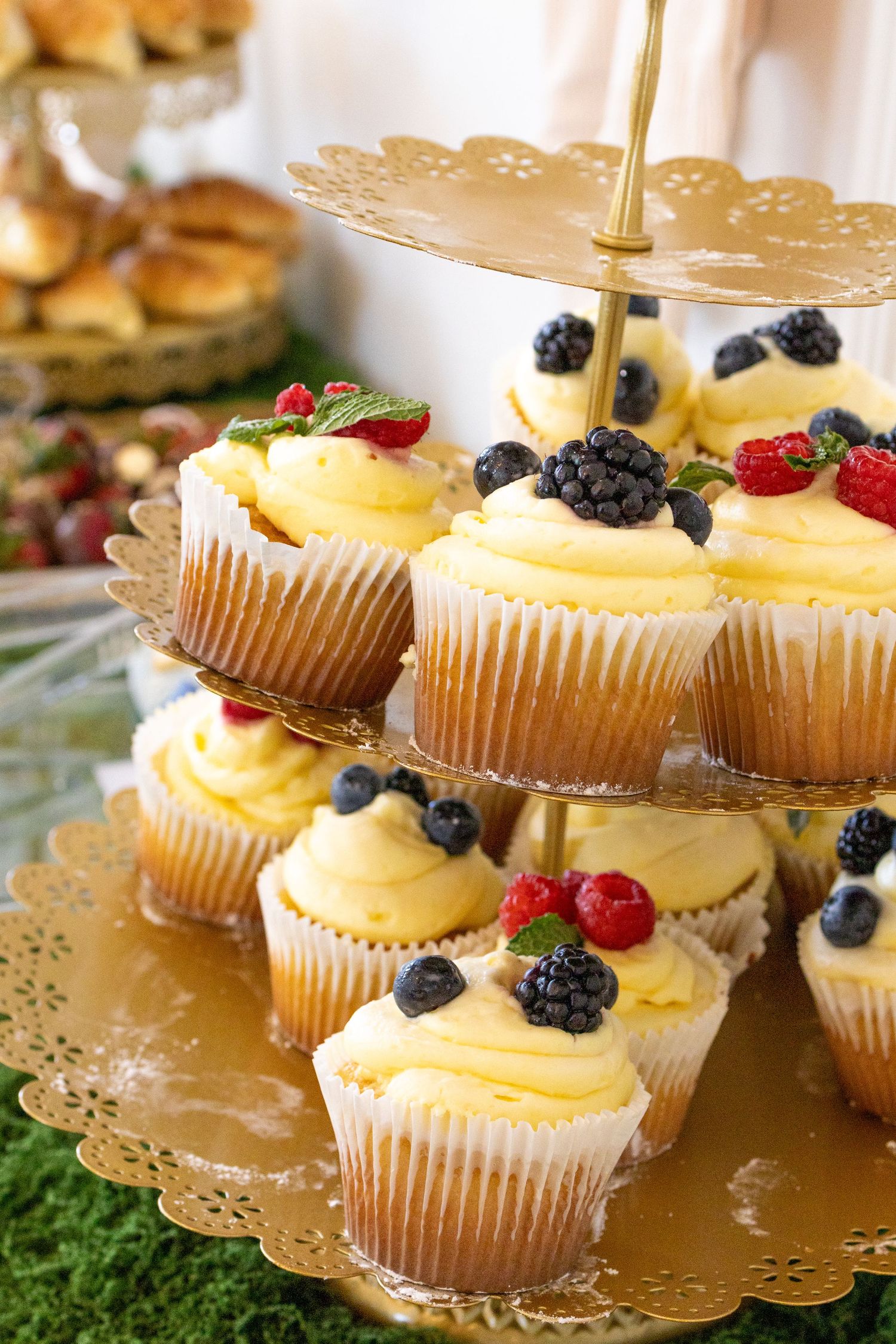 Gold tiered stand displaying vanilla cupcakes topped with yellow frosting, fresh blueberries, raspberries, and blackberries.