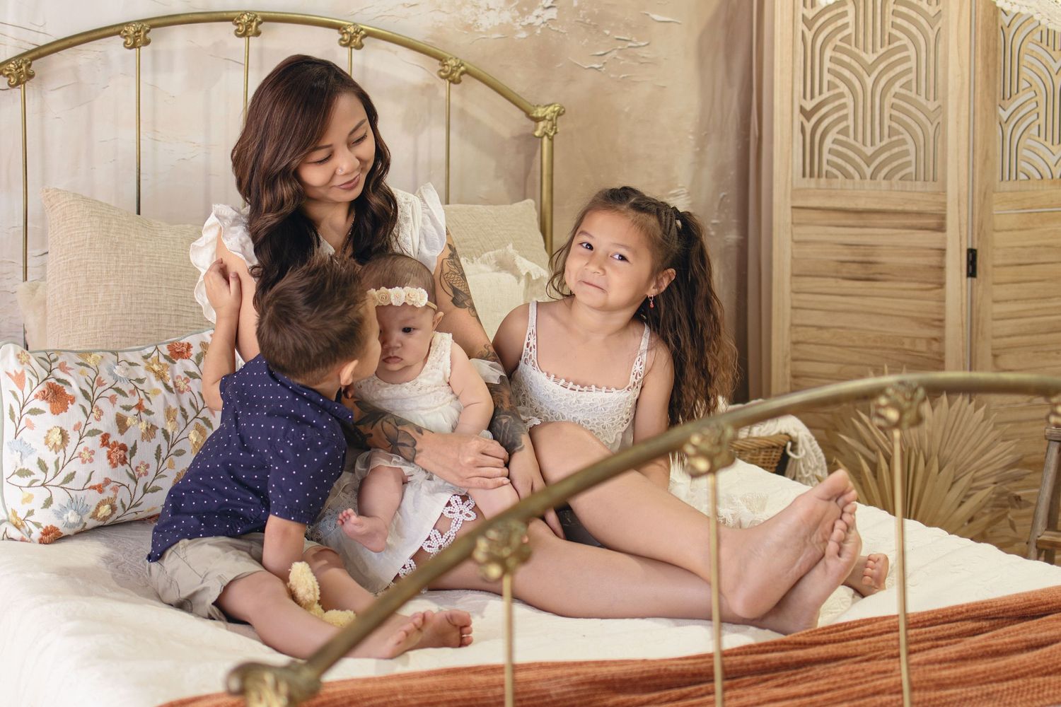 A mother sits on a brass bed with her three young children in a warmly decorated bedroom.