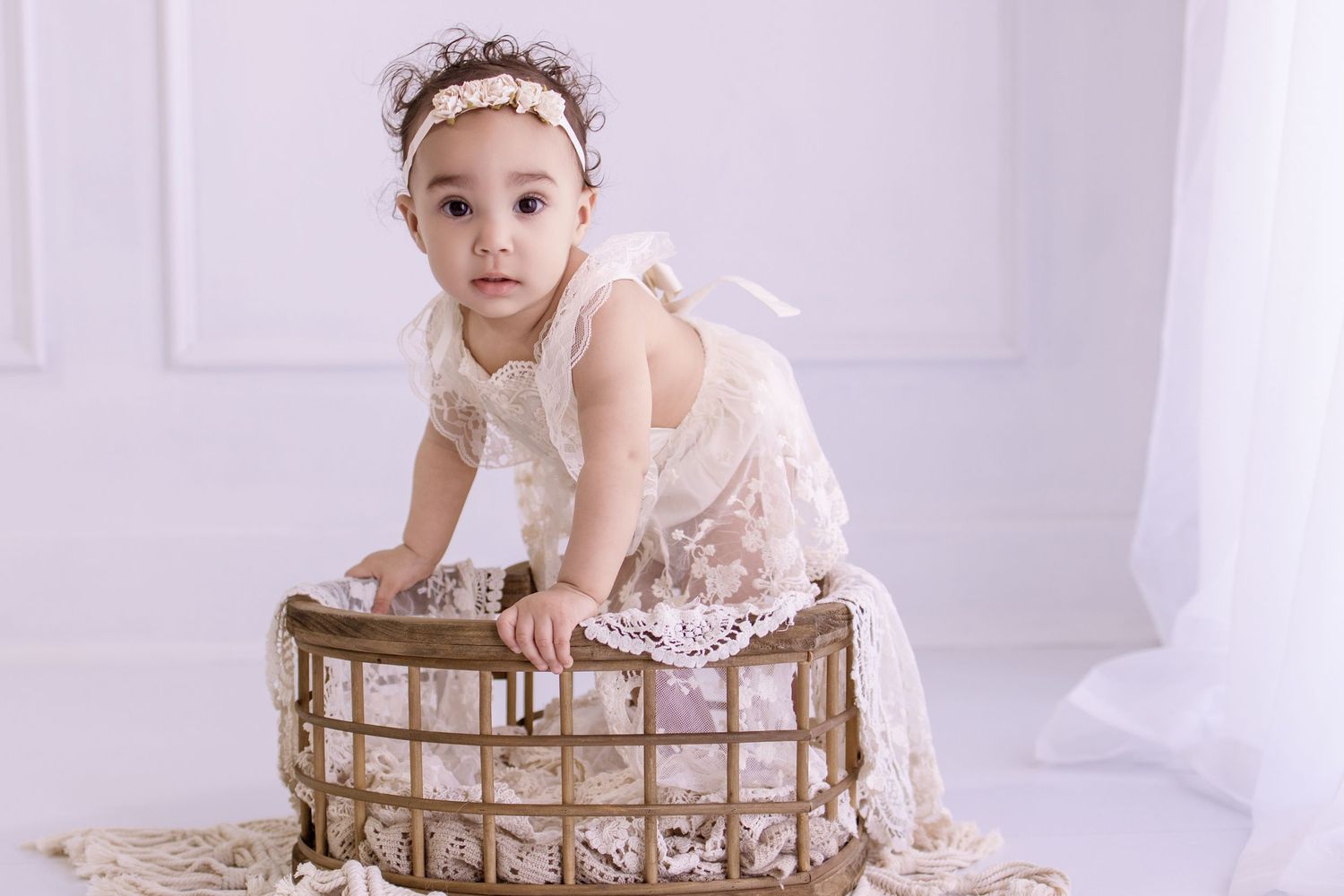 A baby girl in a white lace dress and floral headband poses in a rustic basket in a bright white studio.