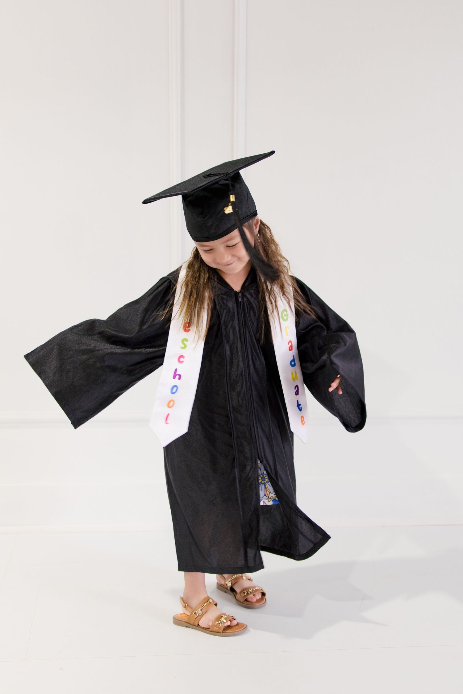 Young girl in black graduation cap and gown with colorful school sash, posing proudly at preschool graduation ceremony.