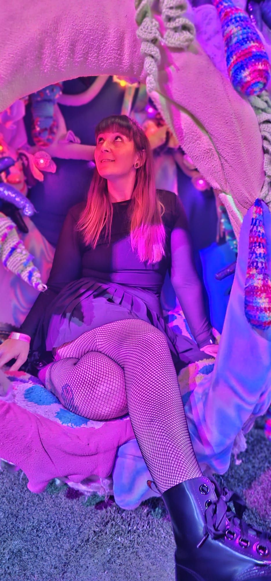 Woman in fishnet tights and black skirt sits in colorful neon-lit art installation at festival.
