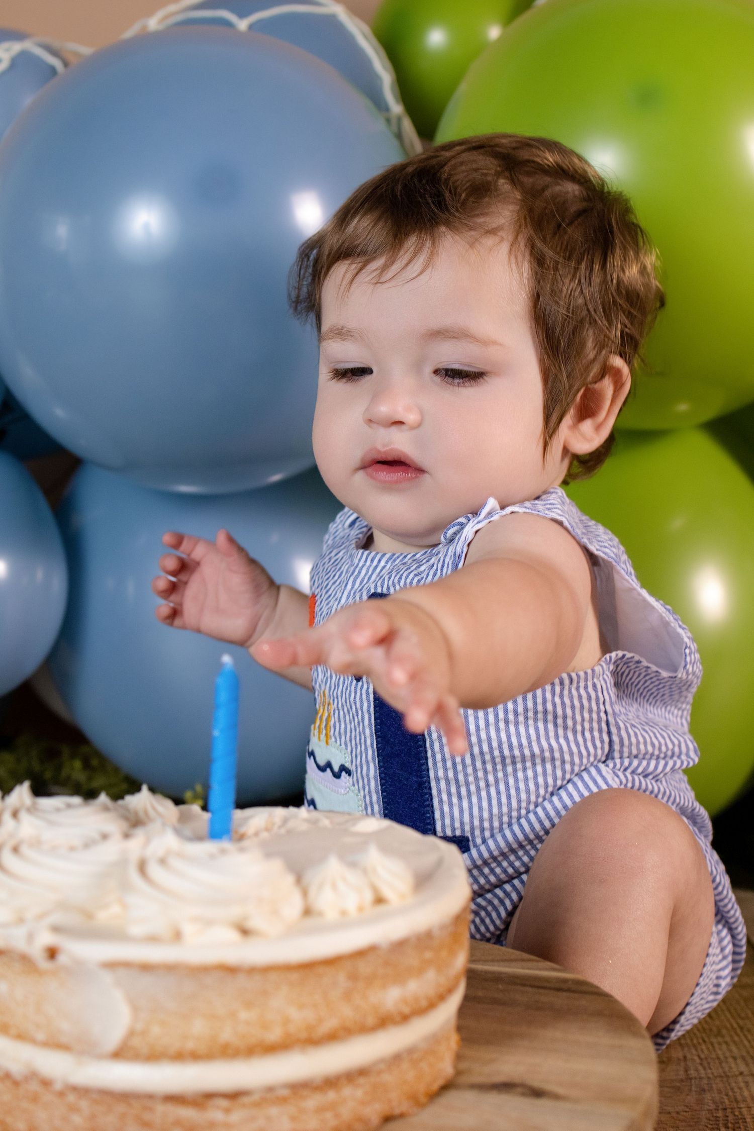 Baby boy reaching for his first birthday cake with a blue candle, surrounded by blue and green balloons.
