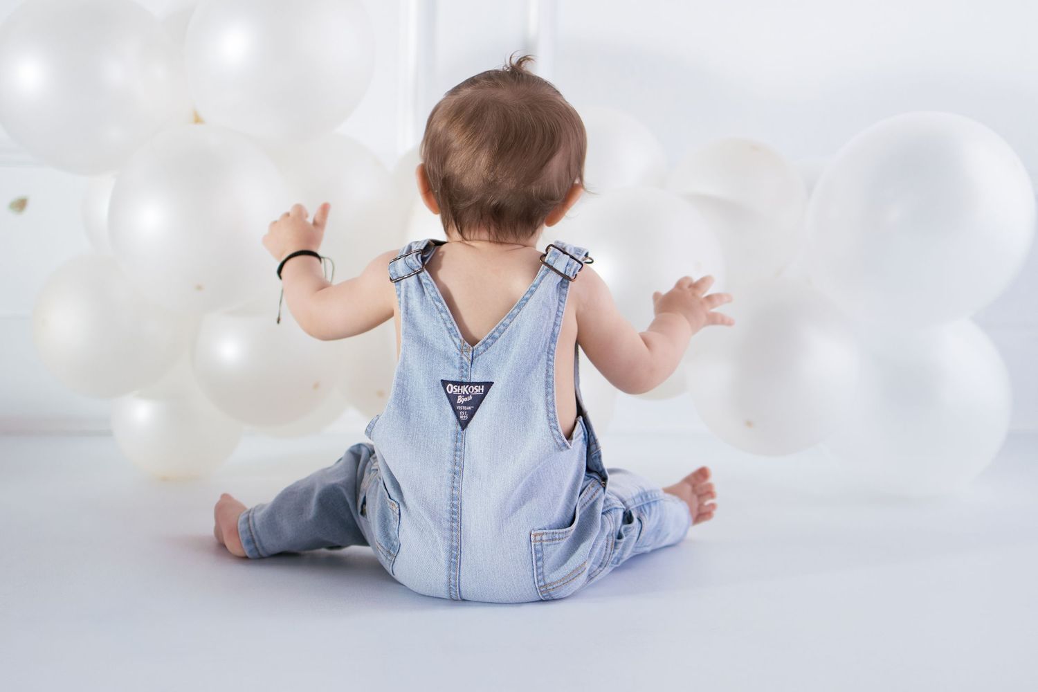 Baby in denim overalls sitting on white floor surrounded by white balloons, viewed from behind.