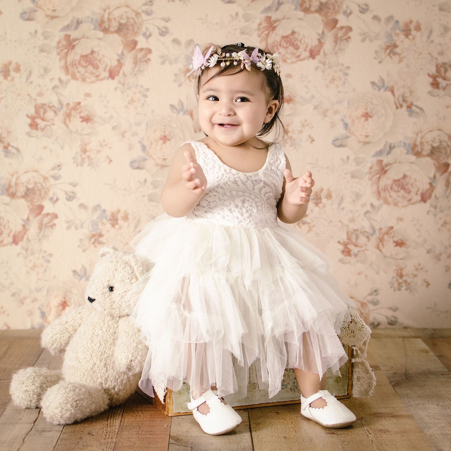 Smiling baby girl in white tulle dress and floral headband poses with teddy bear on wooden floor.