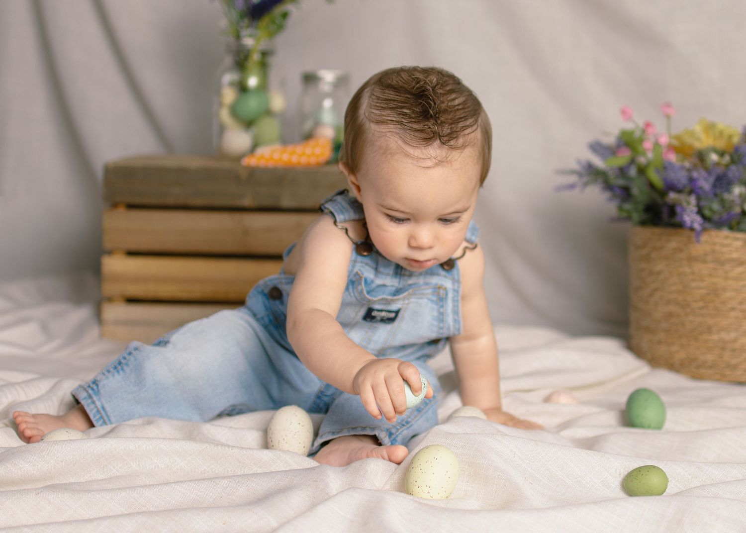 Baby in denim overalls sits on white fabric playing with colorful Easter eggs during a spring photo shoot.