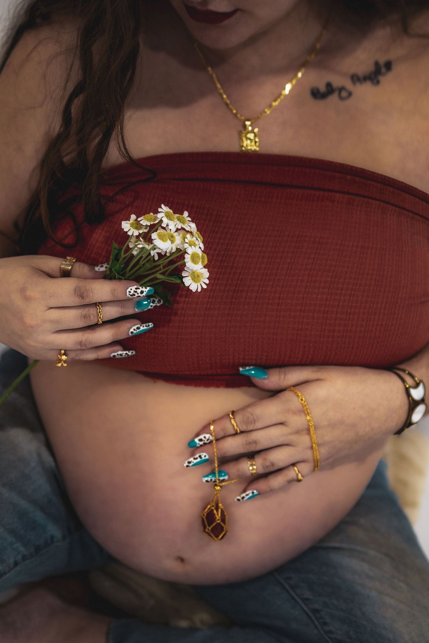 Pregnant woman in red top holding daisies, showcasing decorated nails, gold rings, and necklace while cradling baby bump.