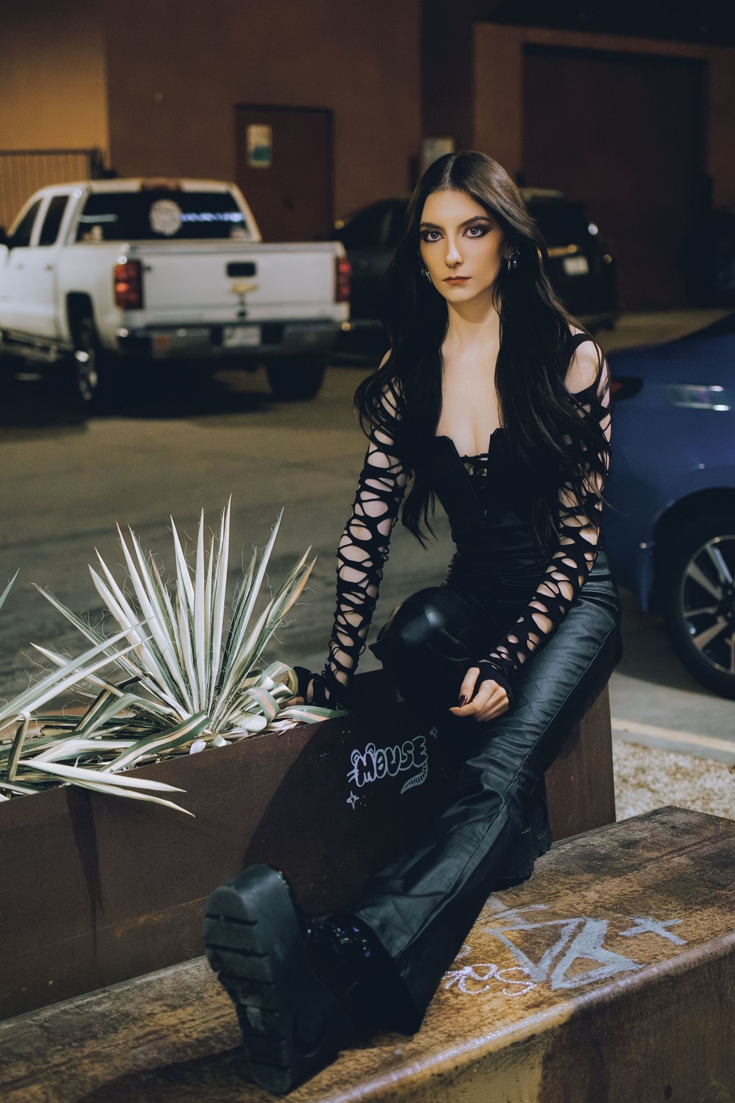 Woman in black cutout sleeves and leather pants posing in urban parking lot at night.