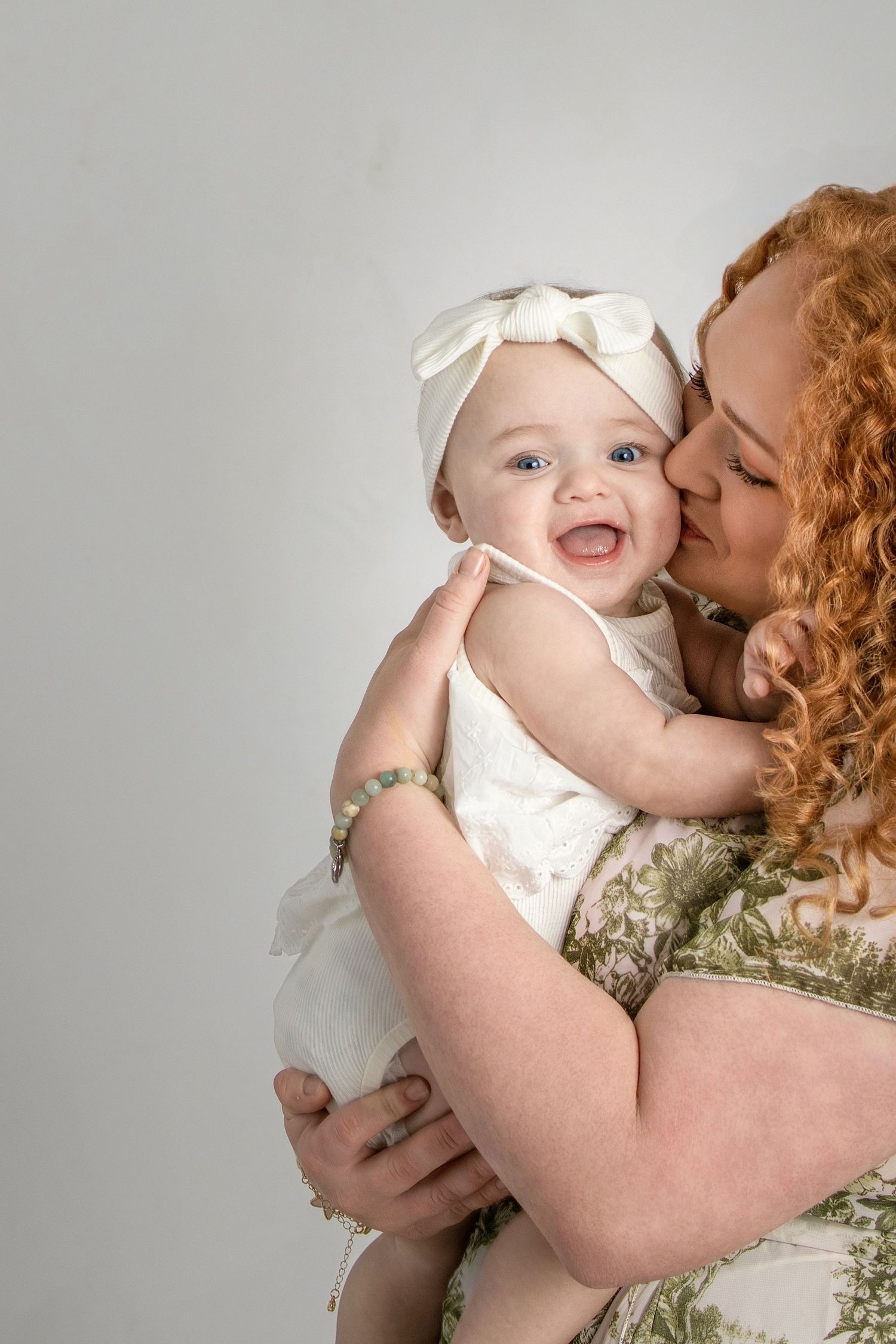 Red-haired mother kissing smiling baby girl in white dress and bow headband.