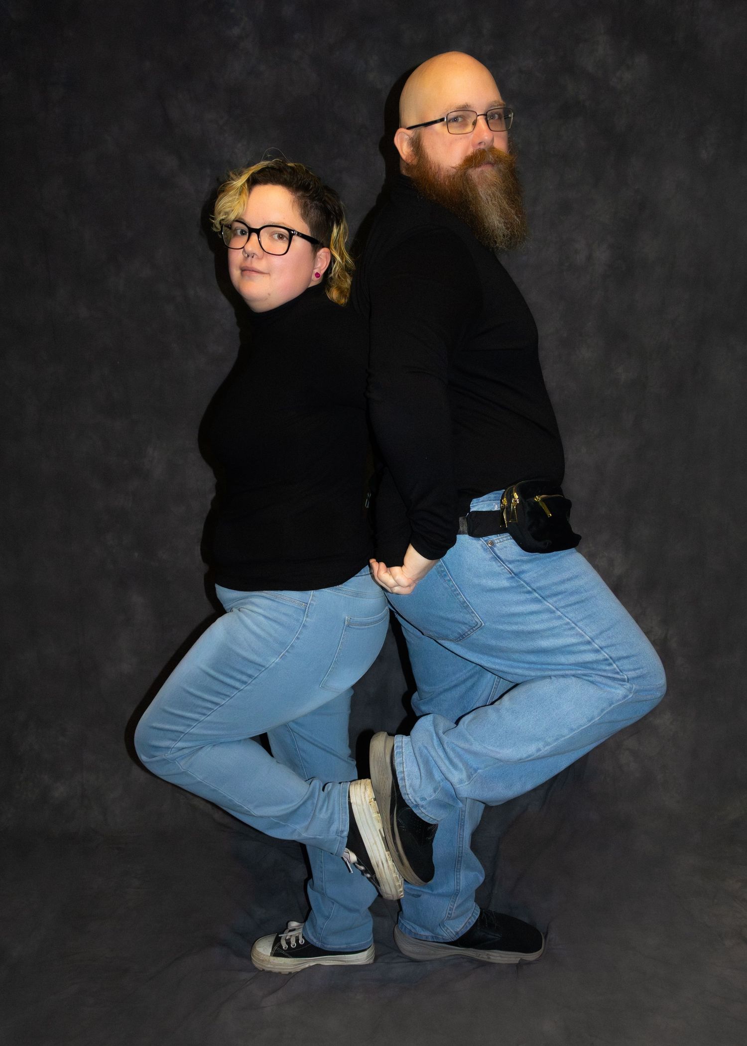 Two people in matching black turtlenecks and blue jeans pose back-to-back in a studio portrait.