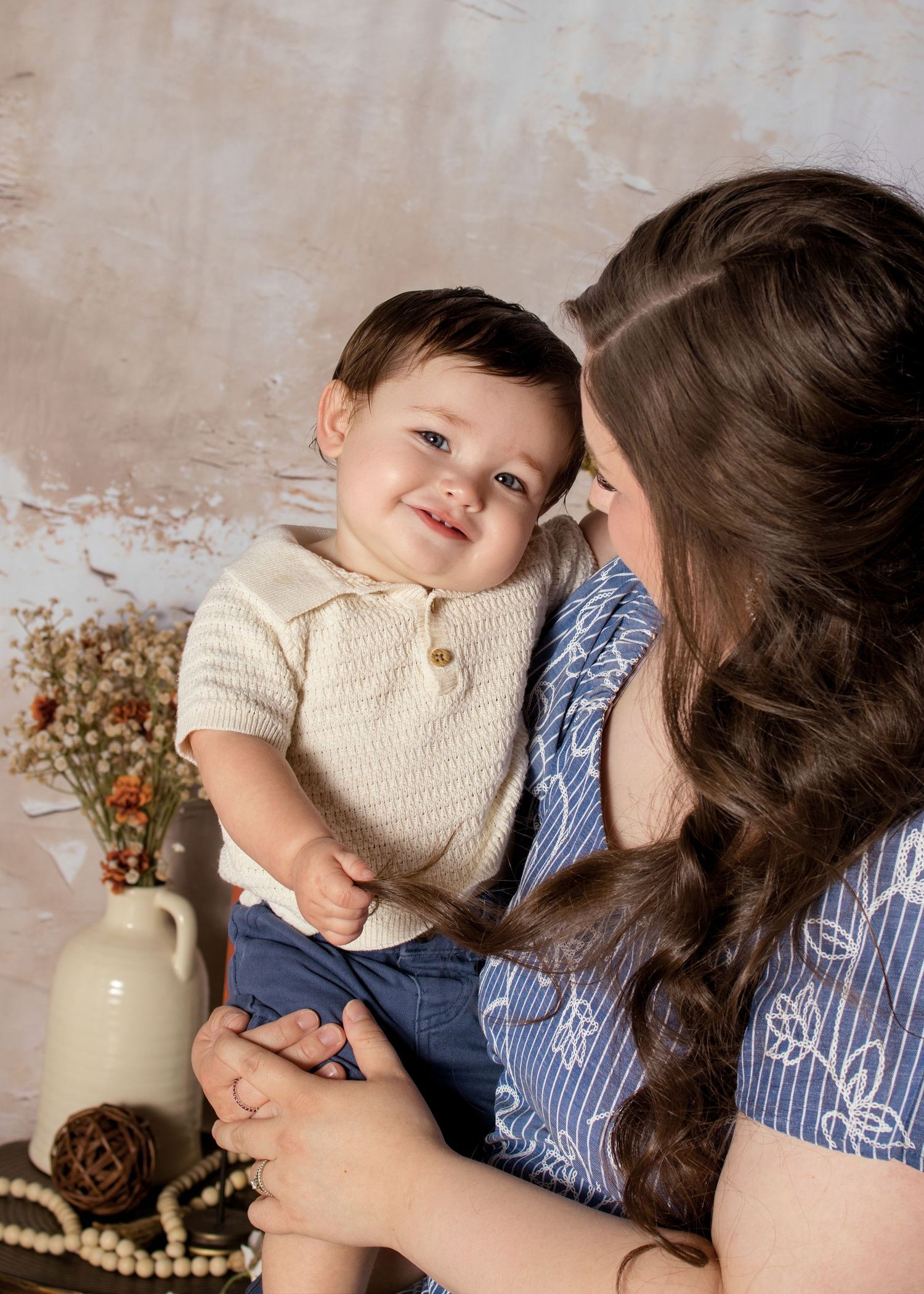 A smiling baby boy in a cream knit top is lovingly held by his dark-haired mother in a blue floral dress.