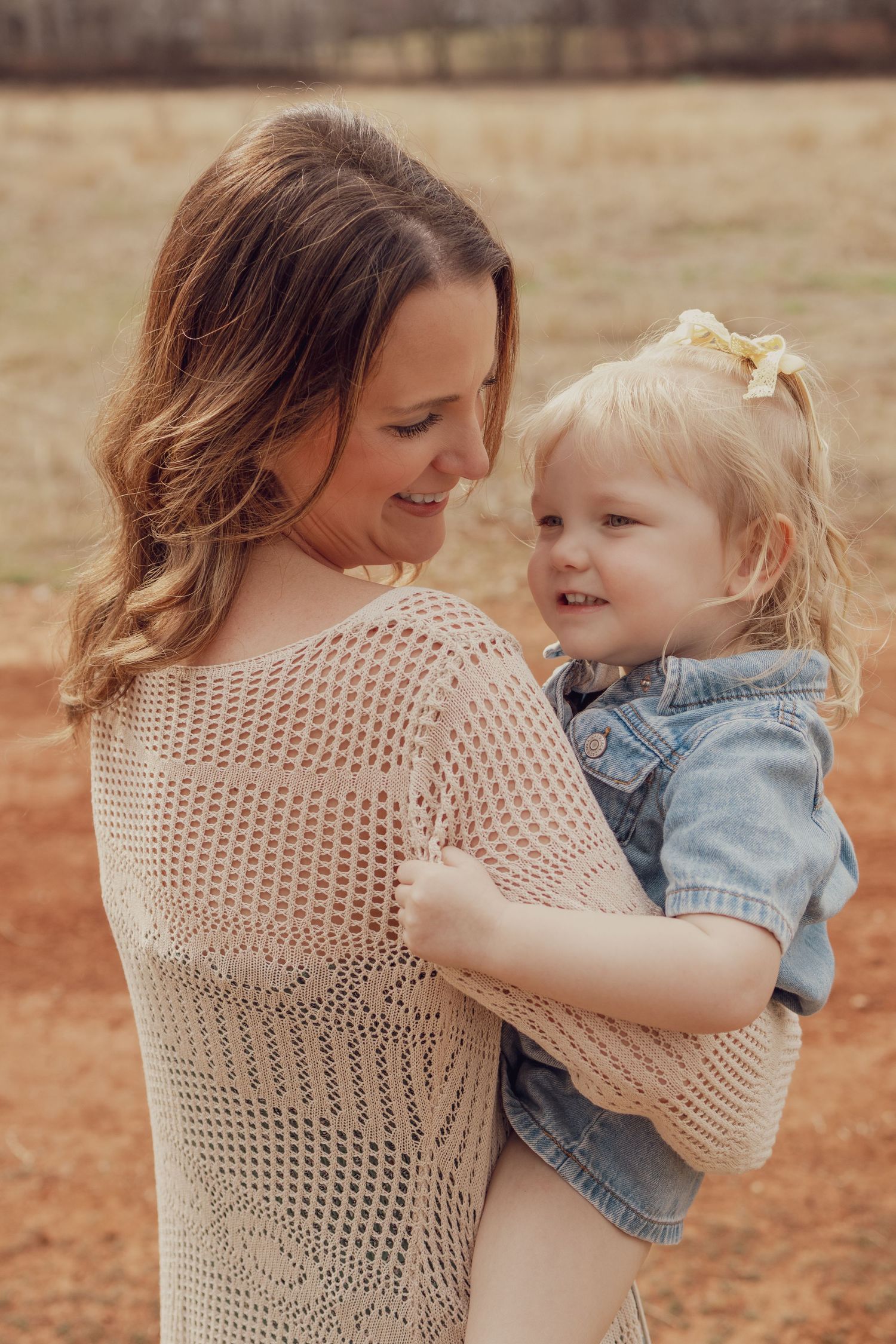 A smiling mother holds her blonde toddler girl in a denim jacket outdoors in a field.