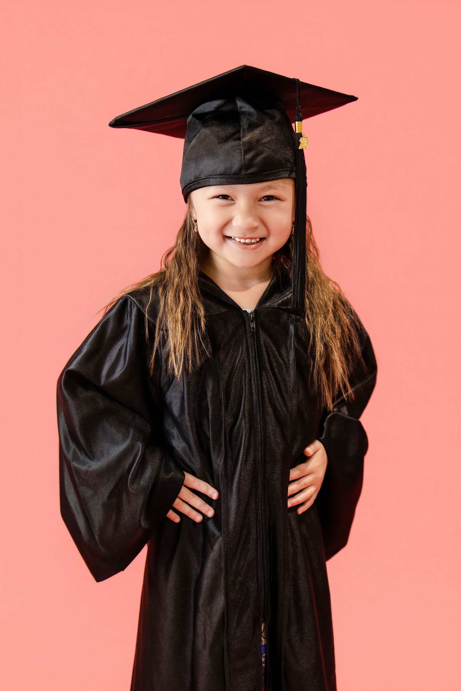 Happy young girl in black graduation cap and gown posing with hands on hips on pink background.
