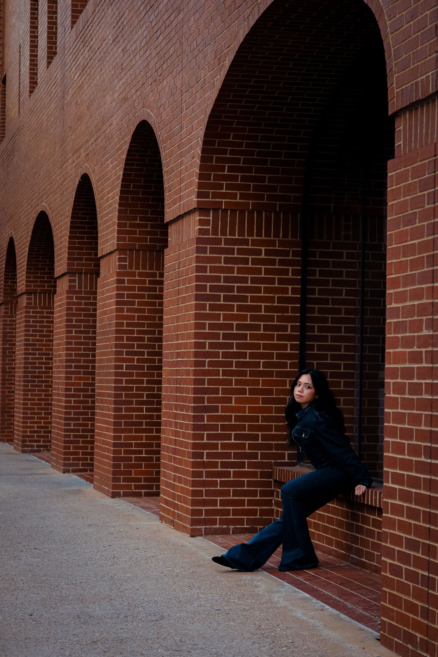 Young Asian woman in dark outfit leaning against red brick arched wall exterior.