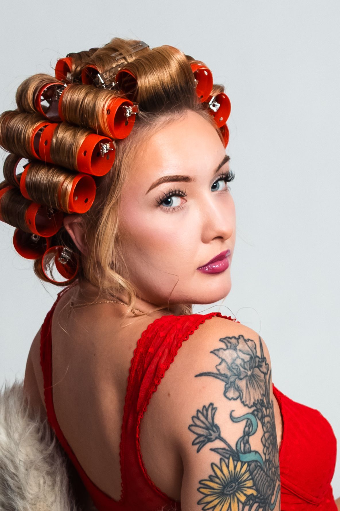 Young woman with orange hair rollers, floral shoulder tattoo, and red dress in studio portrait.