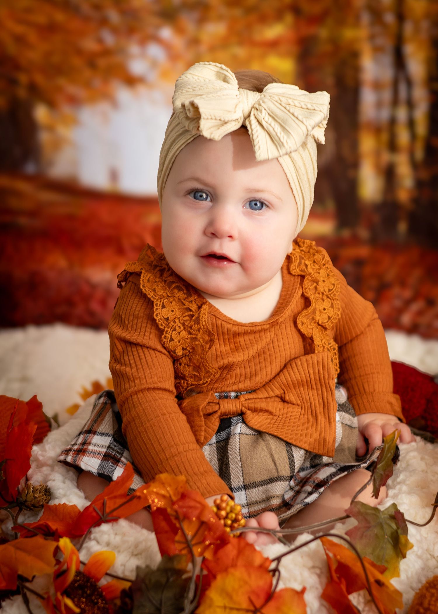 Adorable baby girl in rust orange dress and cream bow headband sits among fall leaves in autumn portrait.