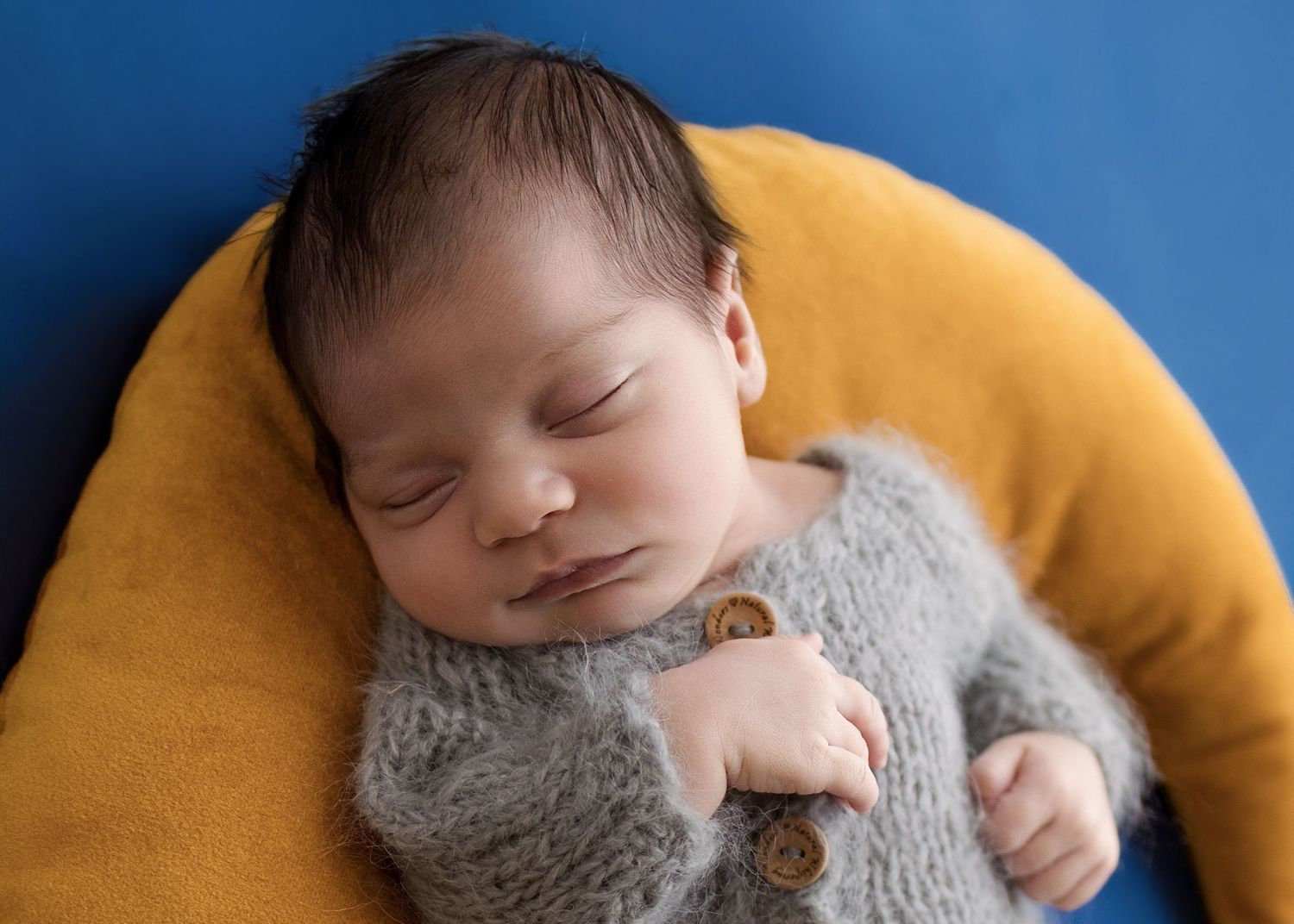 Sleeping newborn baby in gray knit romper with wooden buttons, nestled in yellow bowl on blue background.