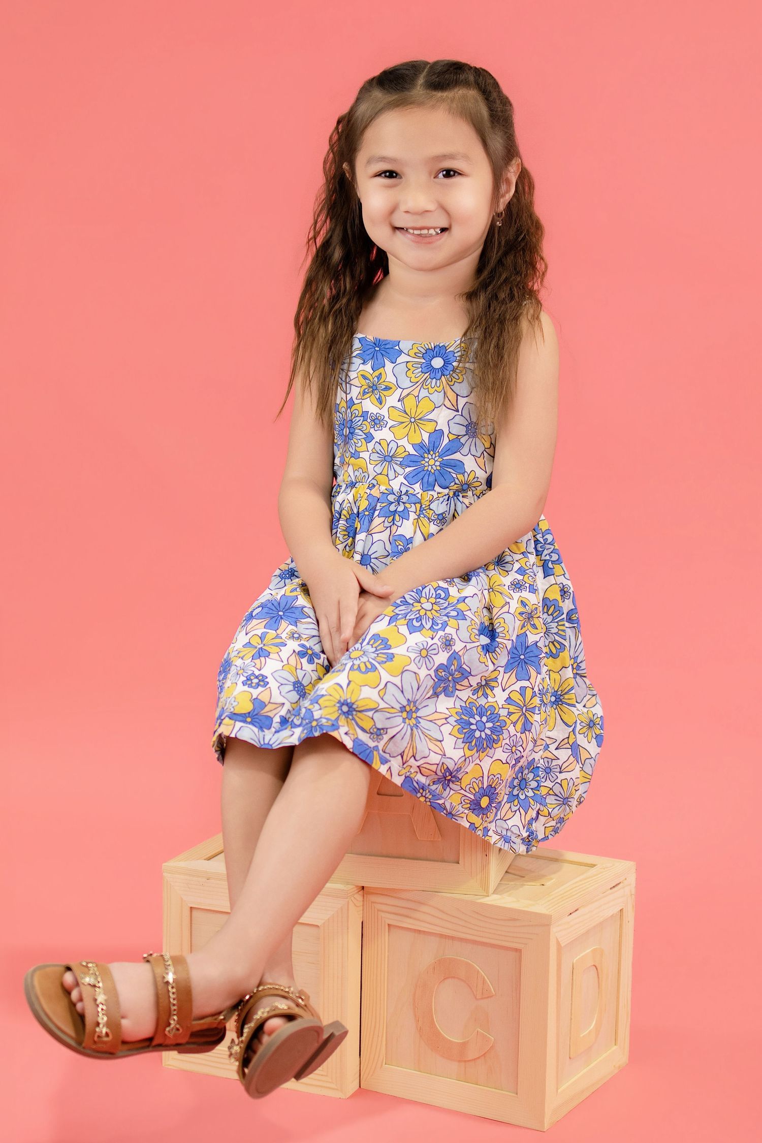 Young girl in floral blue dress sitting on wooden alphabet blocks, smiling, pink background.