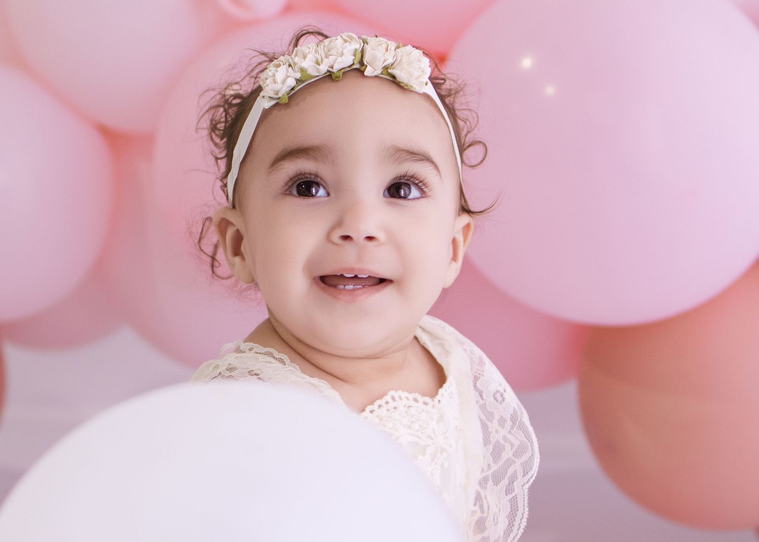 Adorable baby girl in white lace dress and floral headband smiling among pink balloons.
