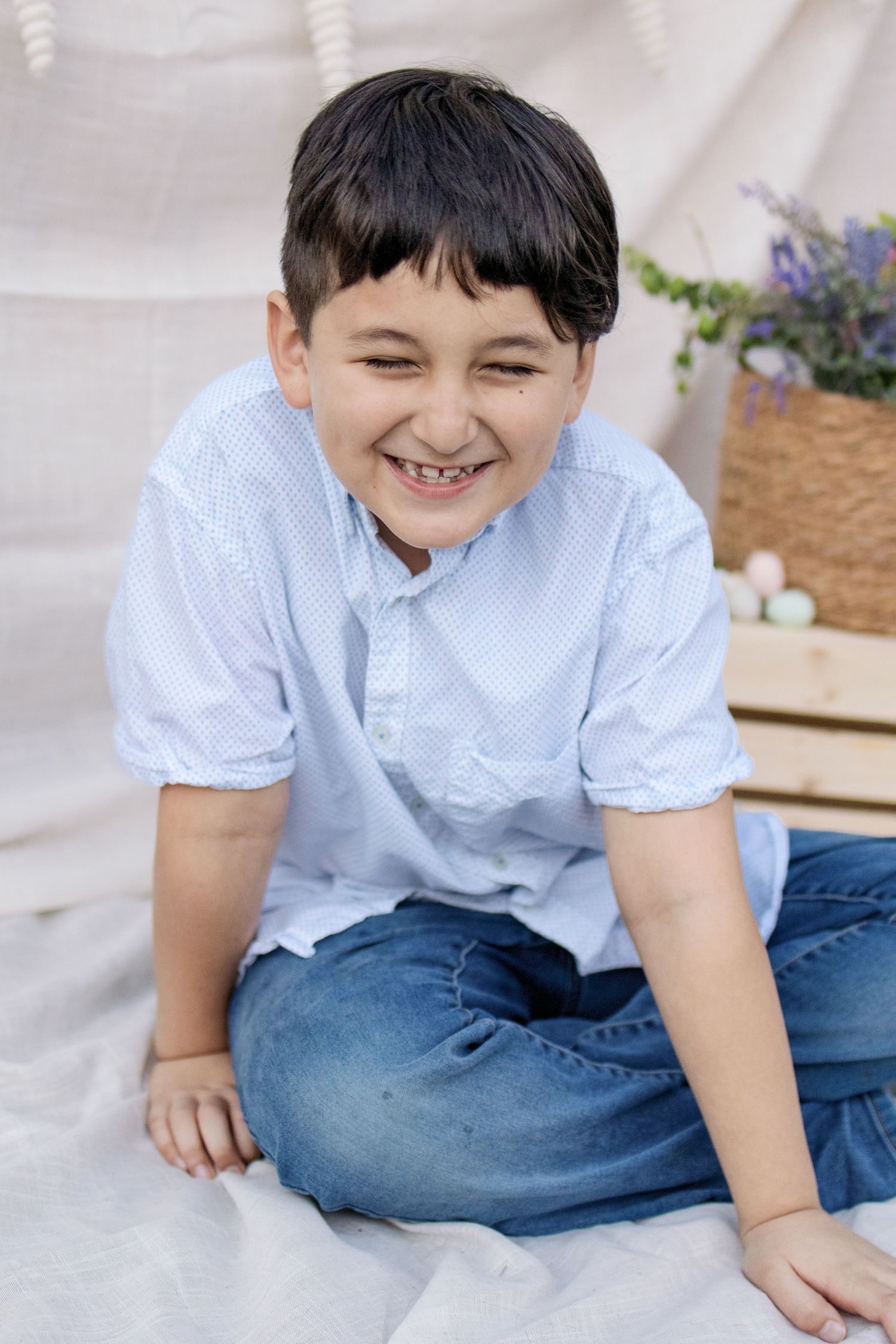 A laughing boy in a light blue shirt and jeans sits cross-legged on a white blanket near Easter eggs.