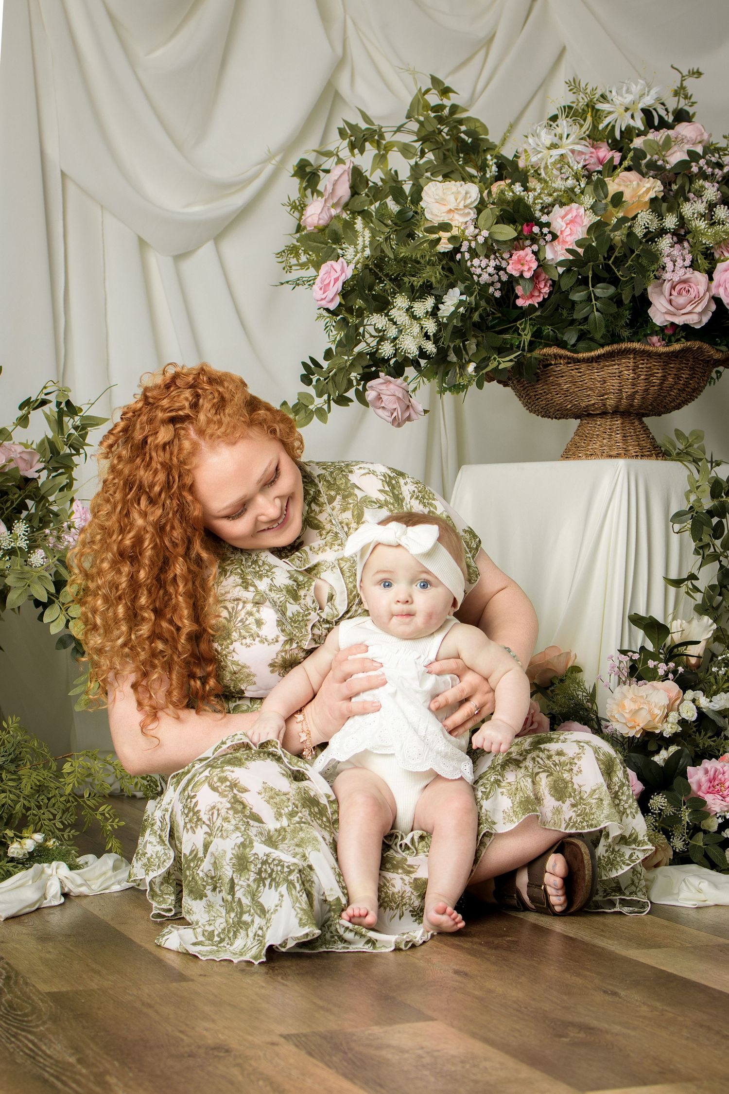 Red-haired mother in floral dress holds baby girl in white outfit among pink roses in studio portrait.