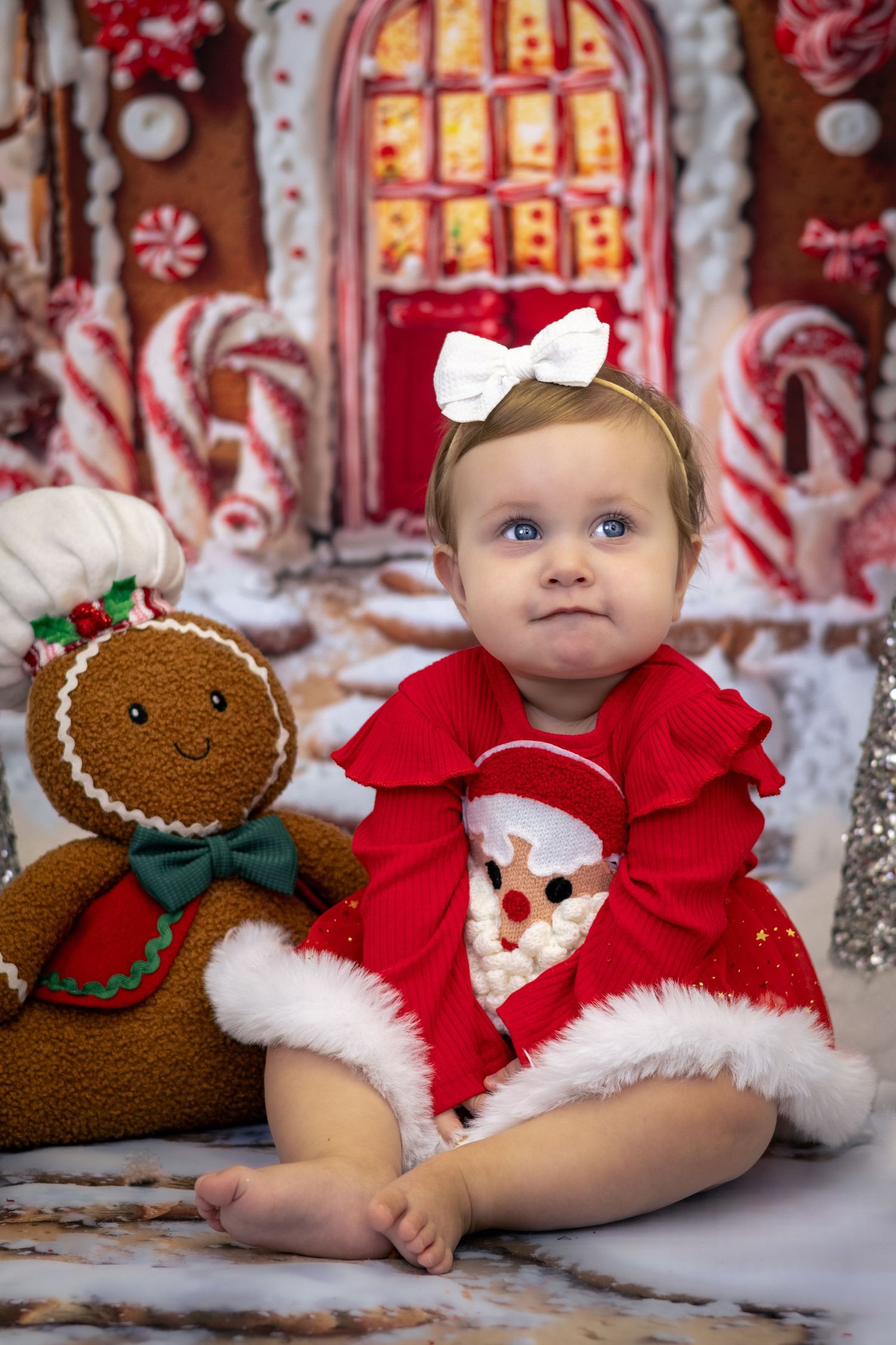 Baby girl in red Santa dress sits beside gingerbread man toy in front of festive candy house backdrop.