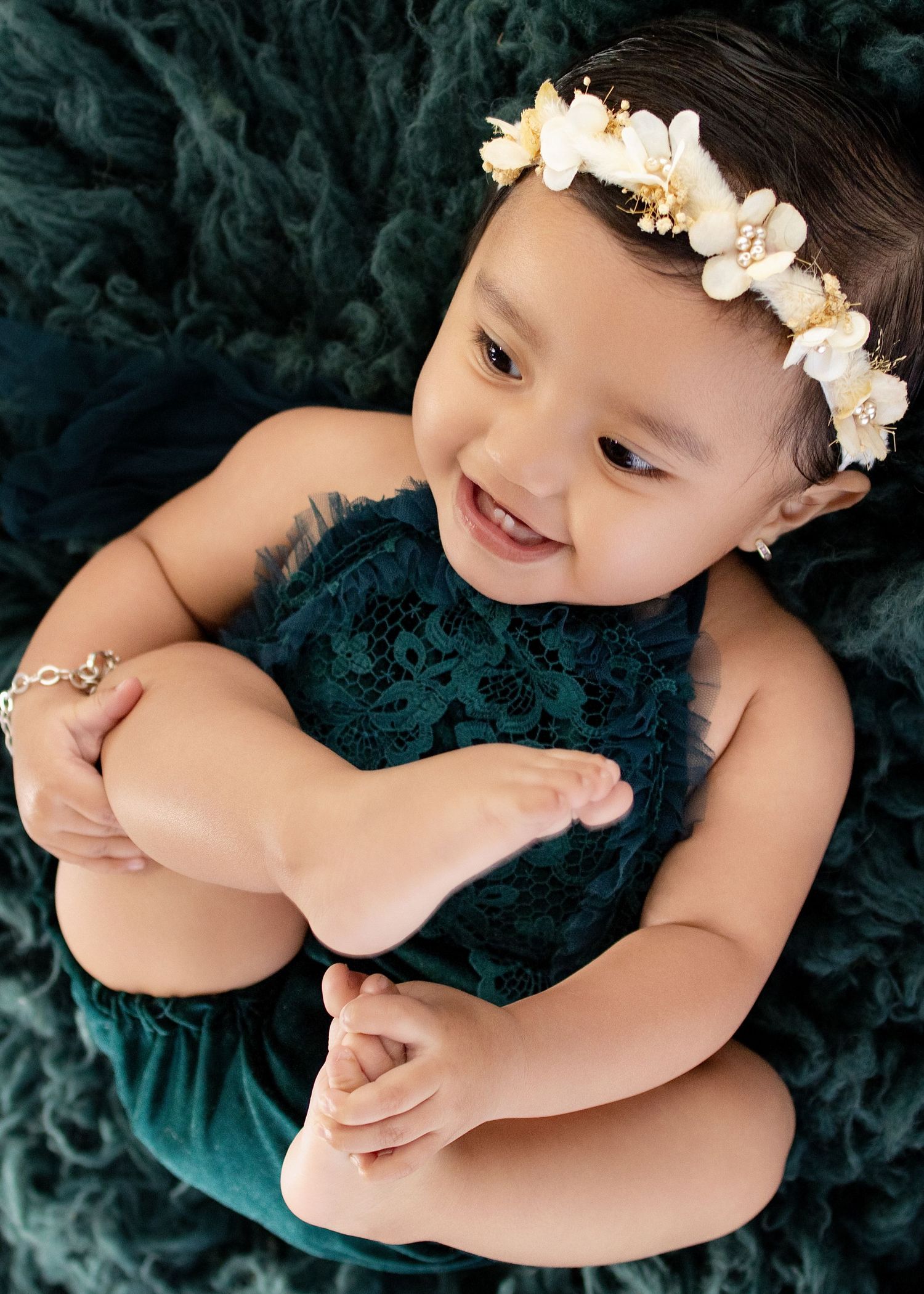 Smiling baby girl in teal lace romper and floral headband, holding her foot on a teal fluffy backdrop.