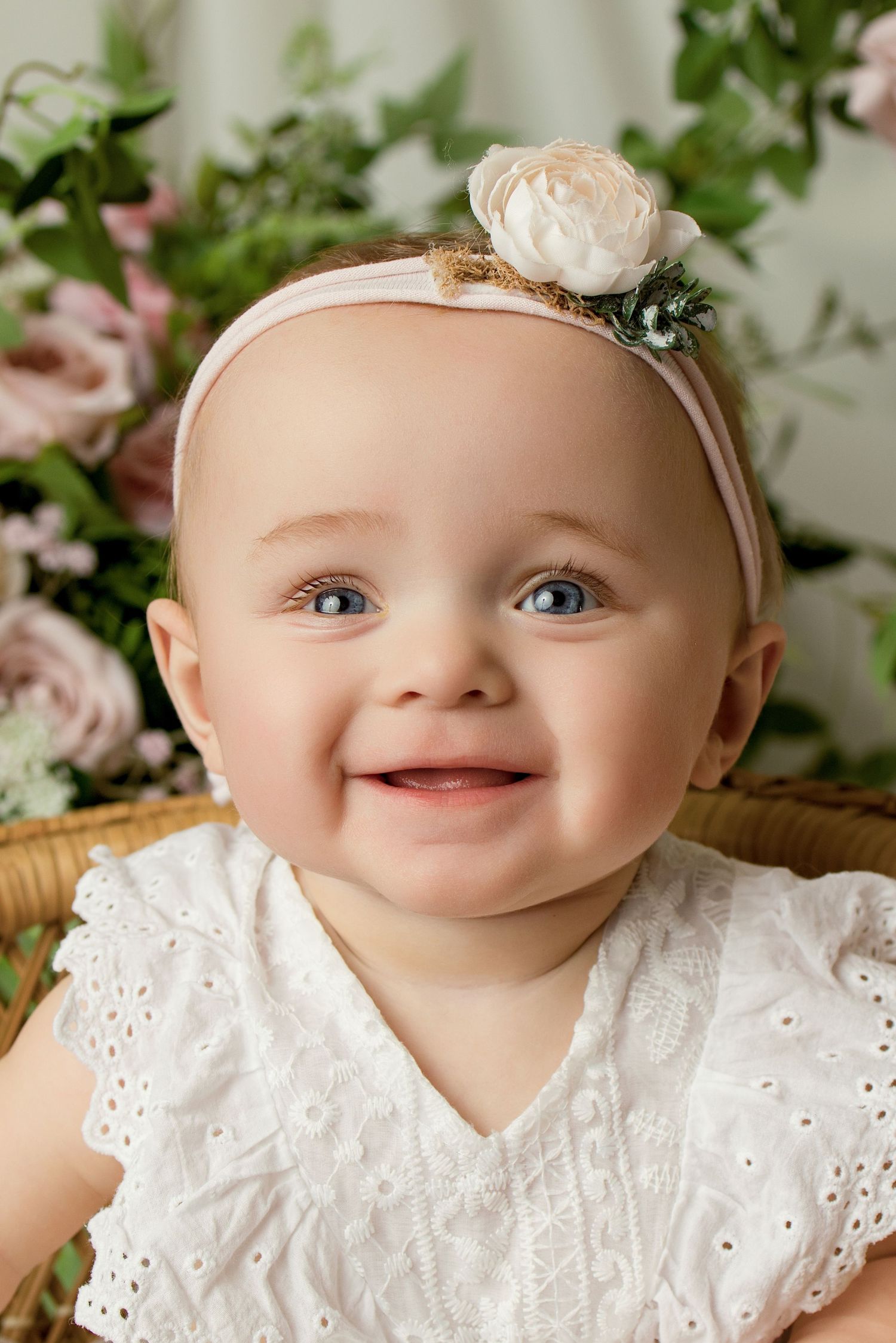 Smiling baby girl with blue eyes wearing a white eyelet dress and floral headband, seated in a wicker chair with pink flowers.