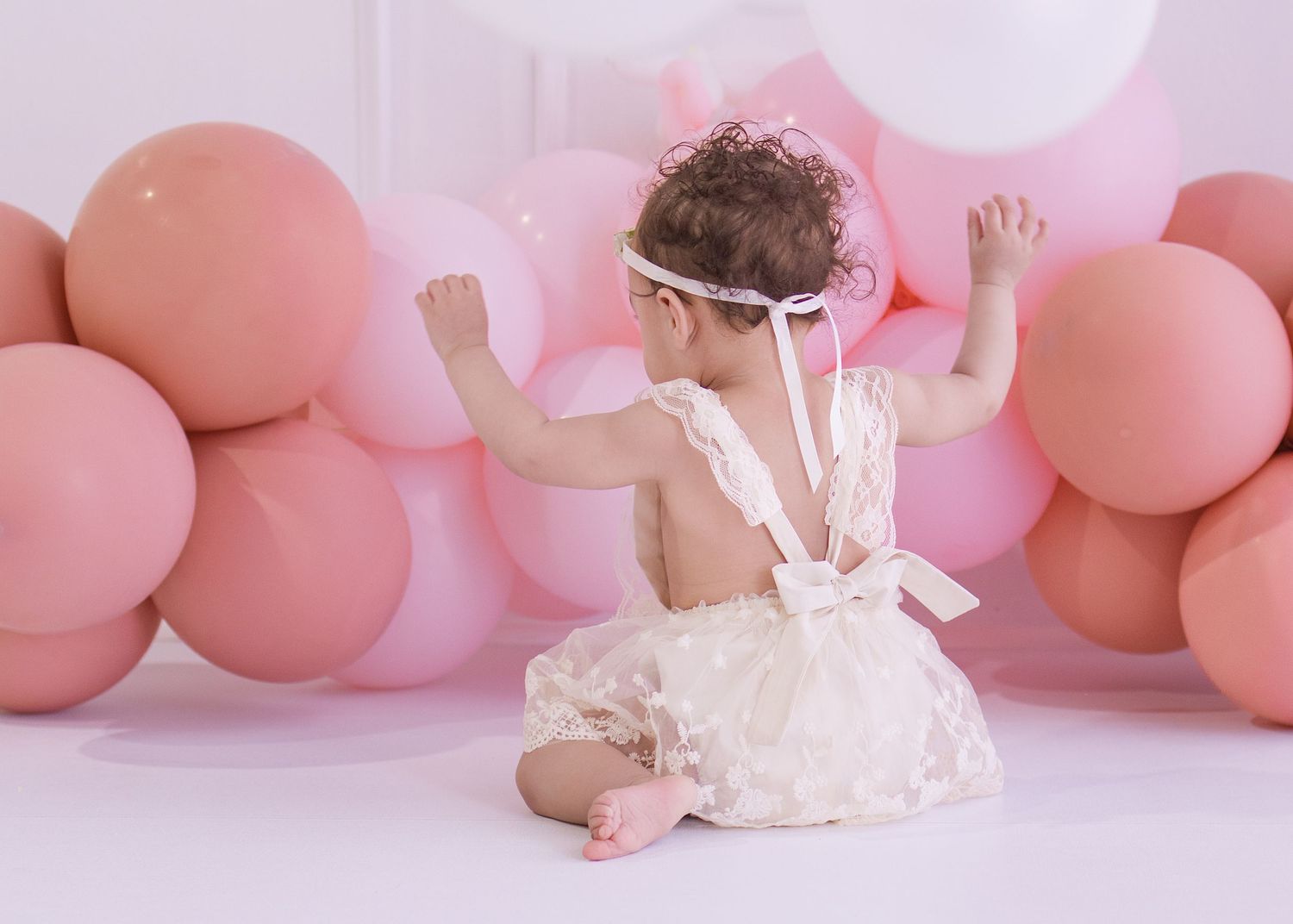 Baby girl in cream dress sits among pink balloons during first birthday cake smash photo session.