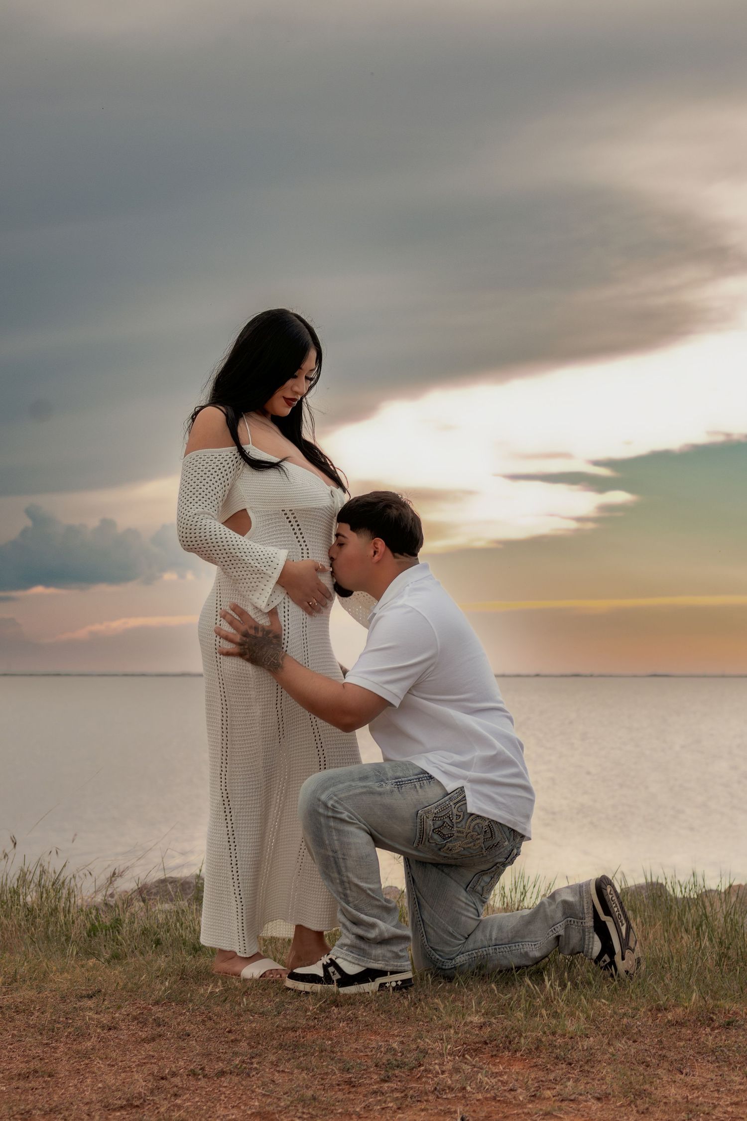 A man kneels kissing a pregnant woman's belly outdoors near water at sunset, both dressed in white.