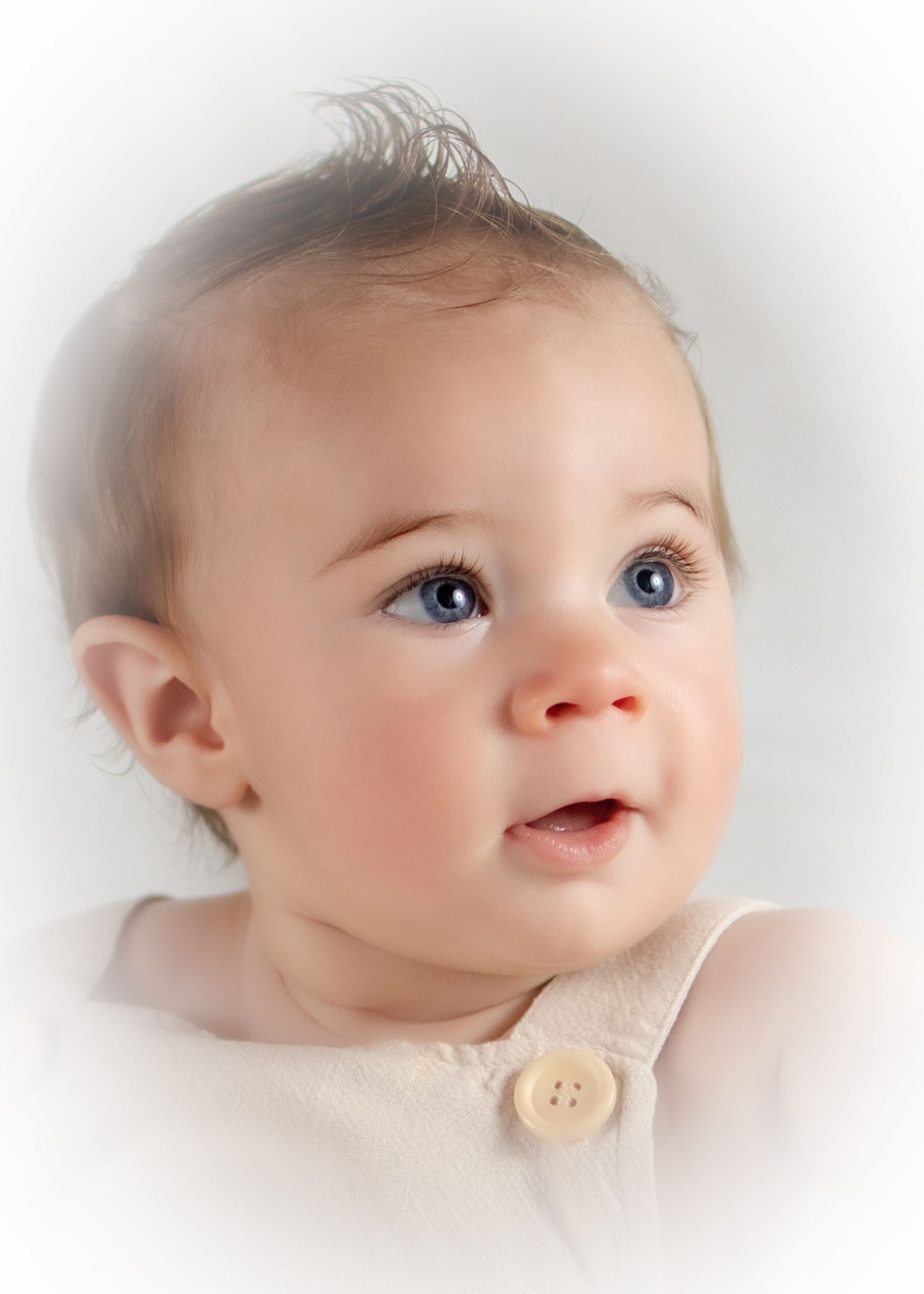 Adorable baby with blue eyes and wispy hair wearing a white outfit, looking up with a curious expression.