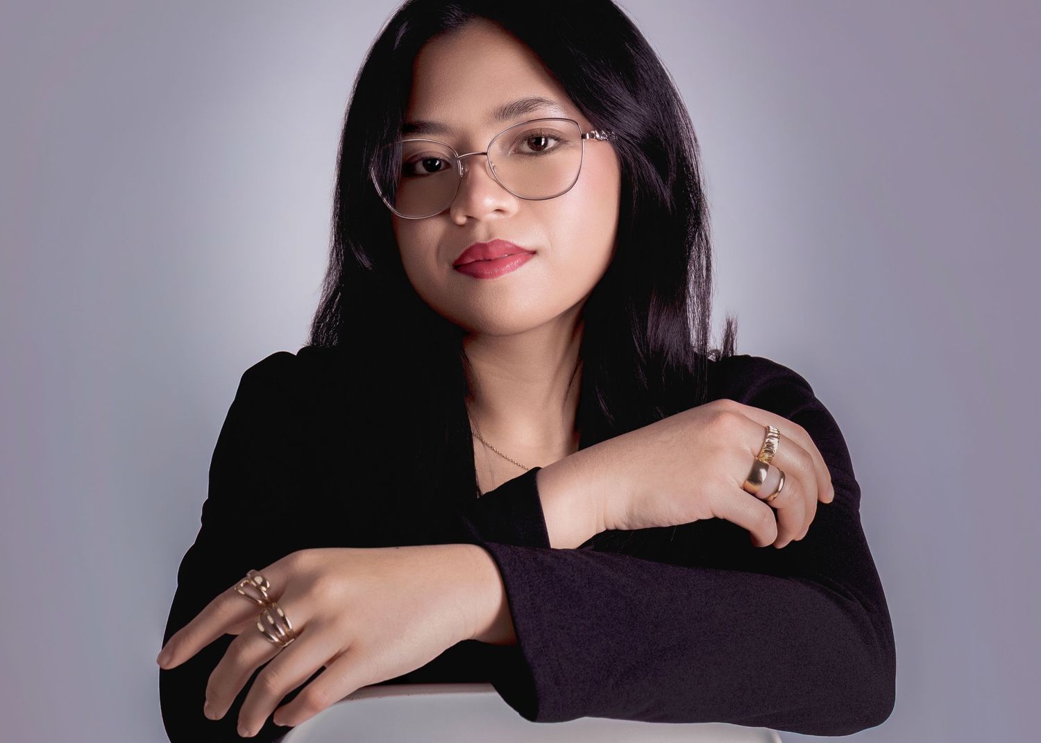 Young woman with glasses and red lipstick posing with arms crossed, wearing a black top and rings.