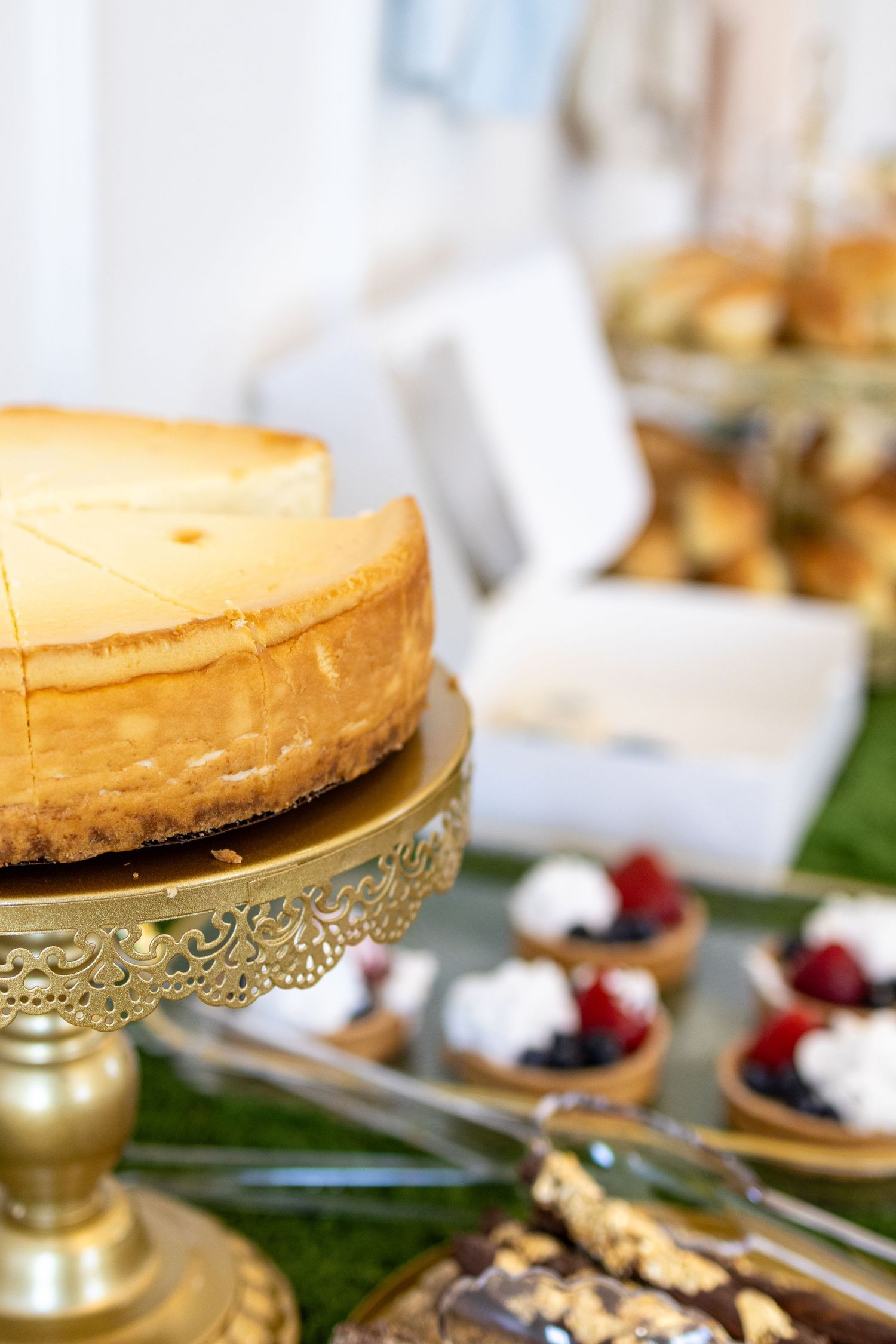 Golden cheesecake on ornate stand with berry tarts and pastries on dessert table.