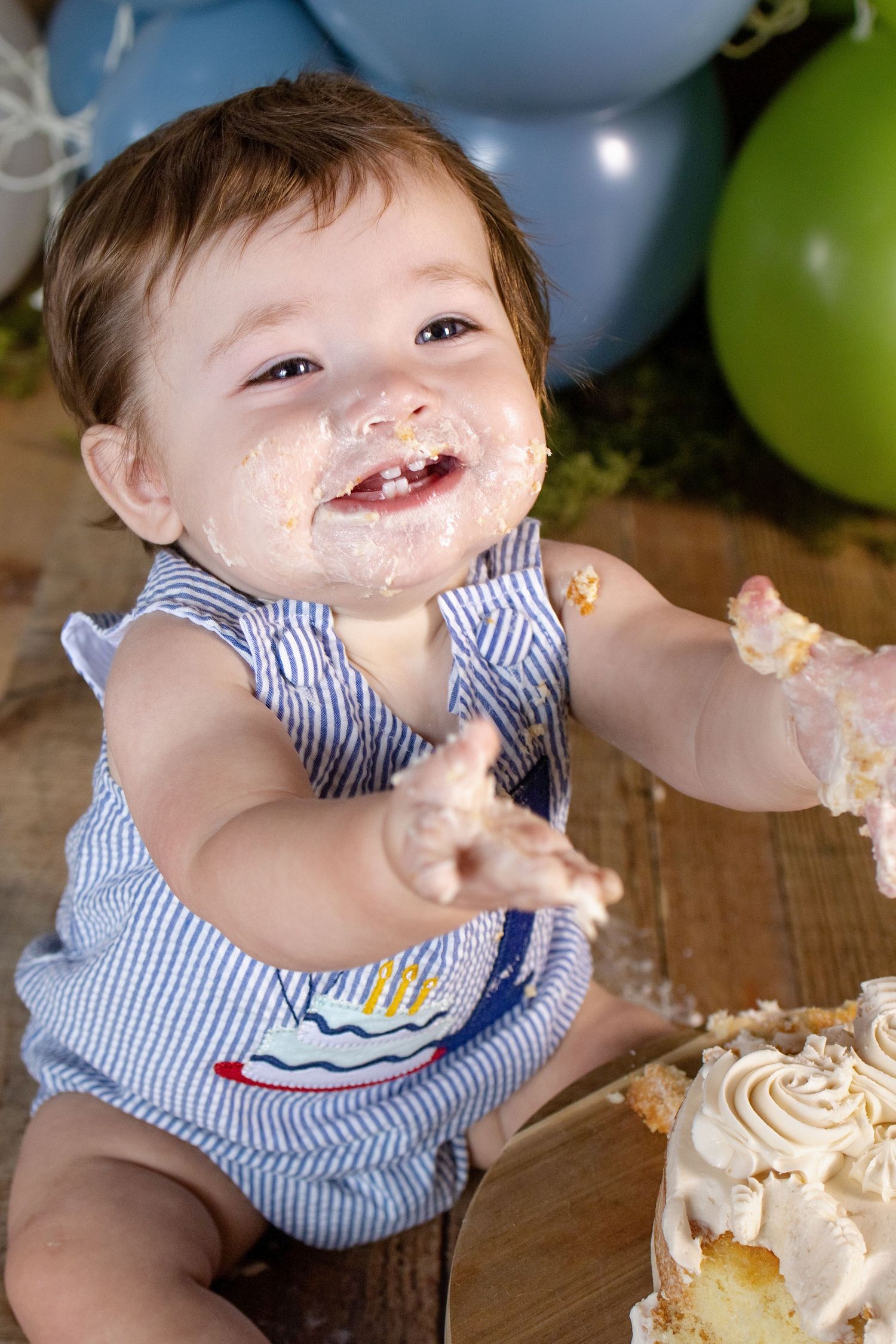 Happy baby with cake-covered face and hands smashing first birthday cake surrounded by blue and green balloons.