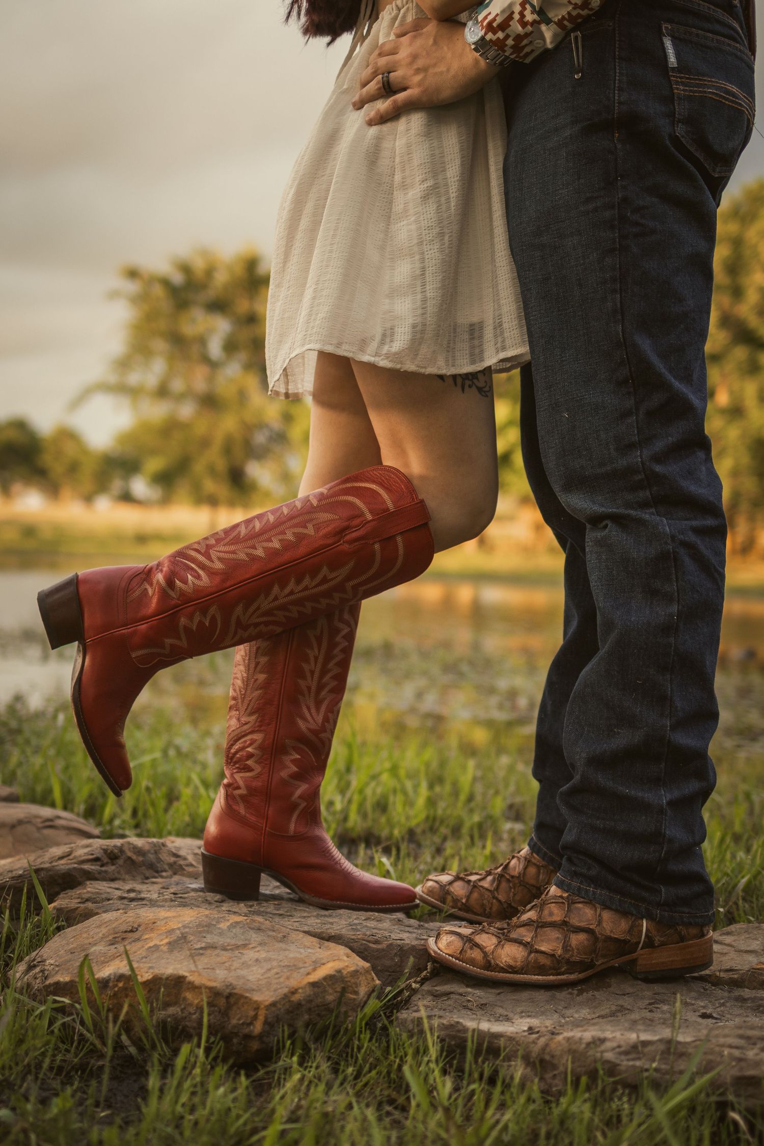 Close-up of a couple standing on rocks, woman in red cowboy boots and white dress, man in jeans and sandals outdoors.