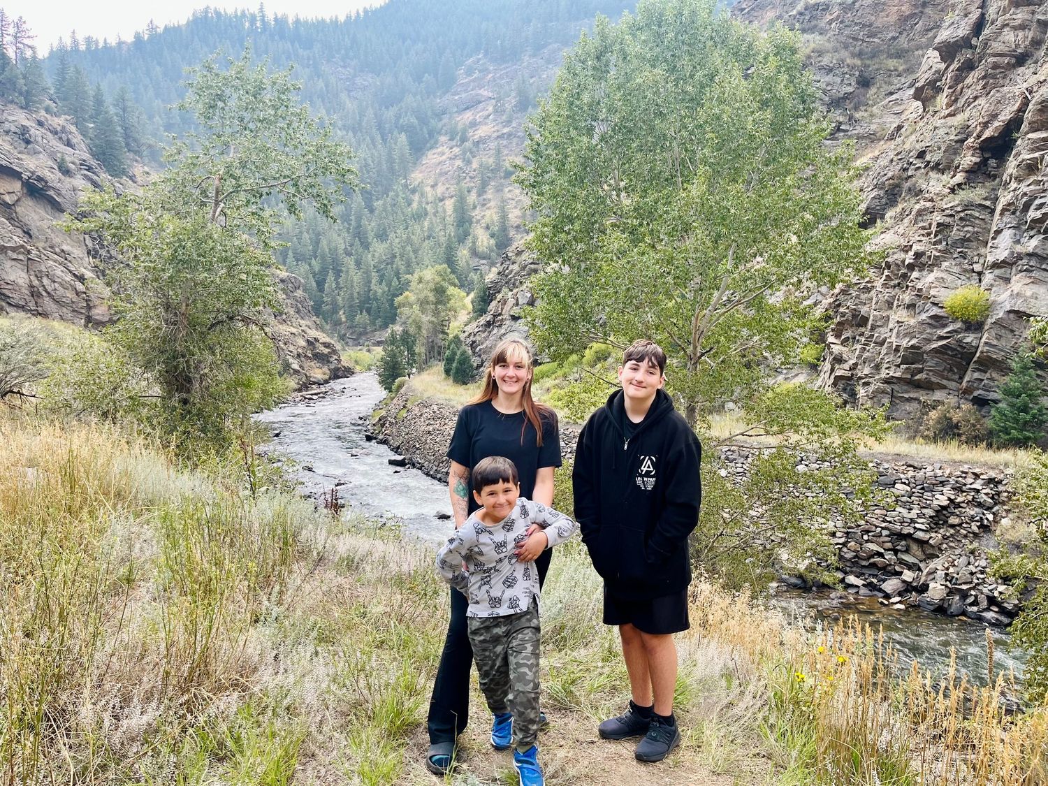 Three people pose near a rocky mountain river canyon with pine trees and rugged cliffs in the background.