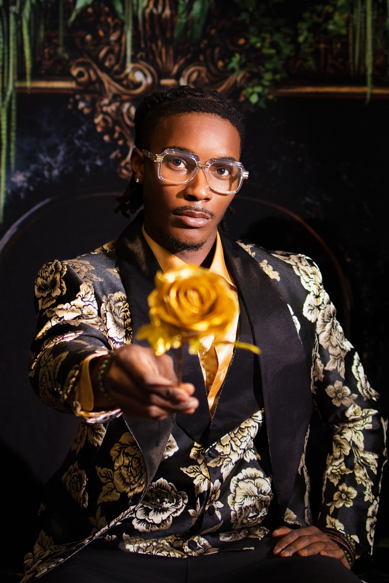 Elegant man in floral gold blazer holds golden rose toward camera while seated in ornate dark interior.