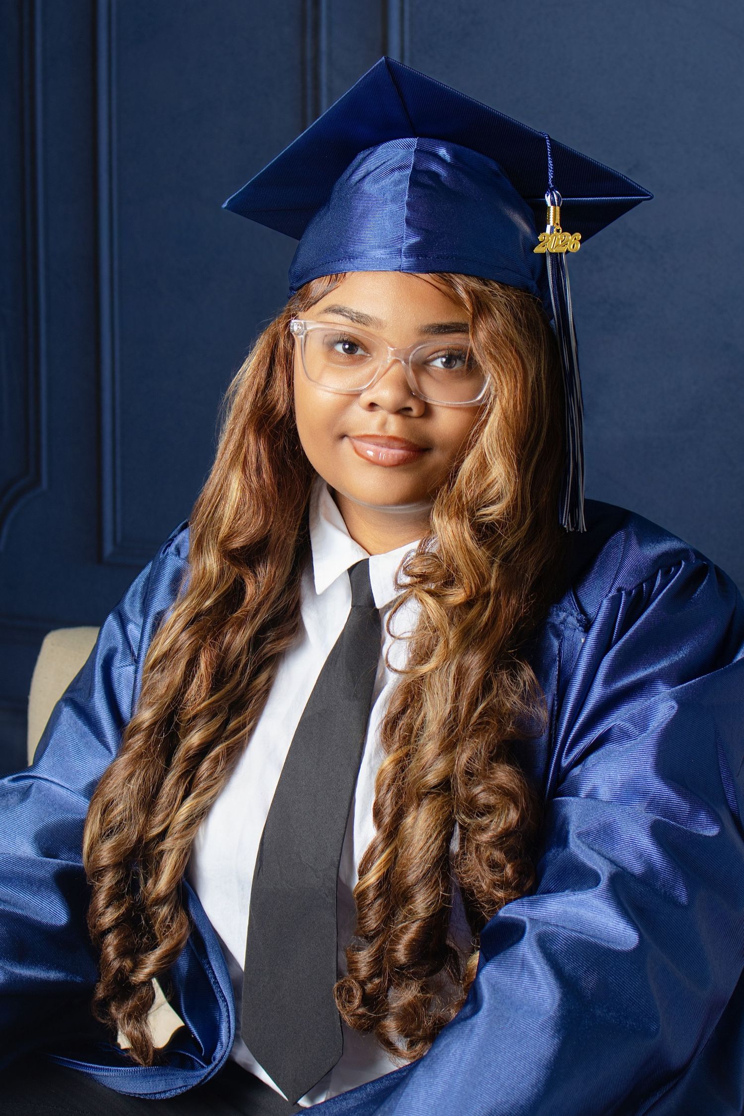 Young woman in blue graduation cap and gown with glasses and black tie posing for portrait photo.