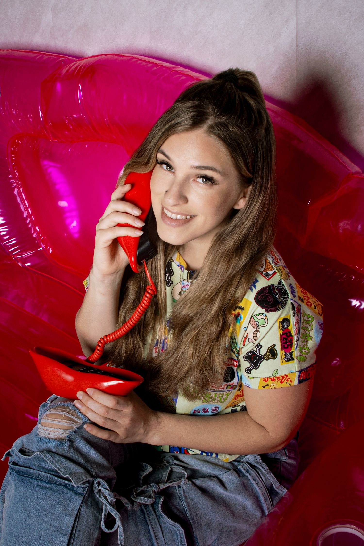 Young woman smiling while holding a red vintage telephone, sitting on a pink inflatable chair.