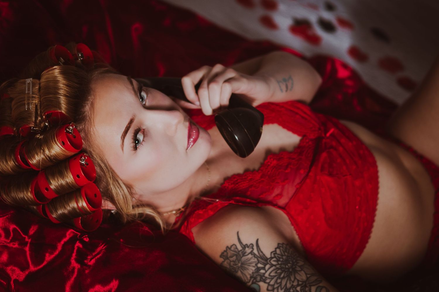 Woman in red lingerie lying down, holding a black phone, with tattoos and red floral decor around her.