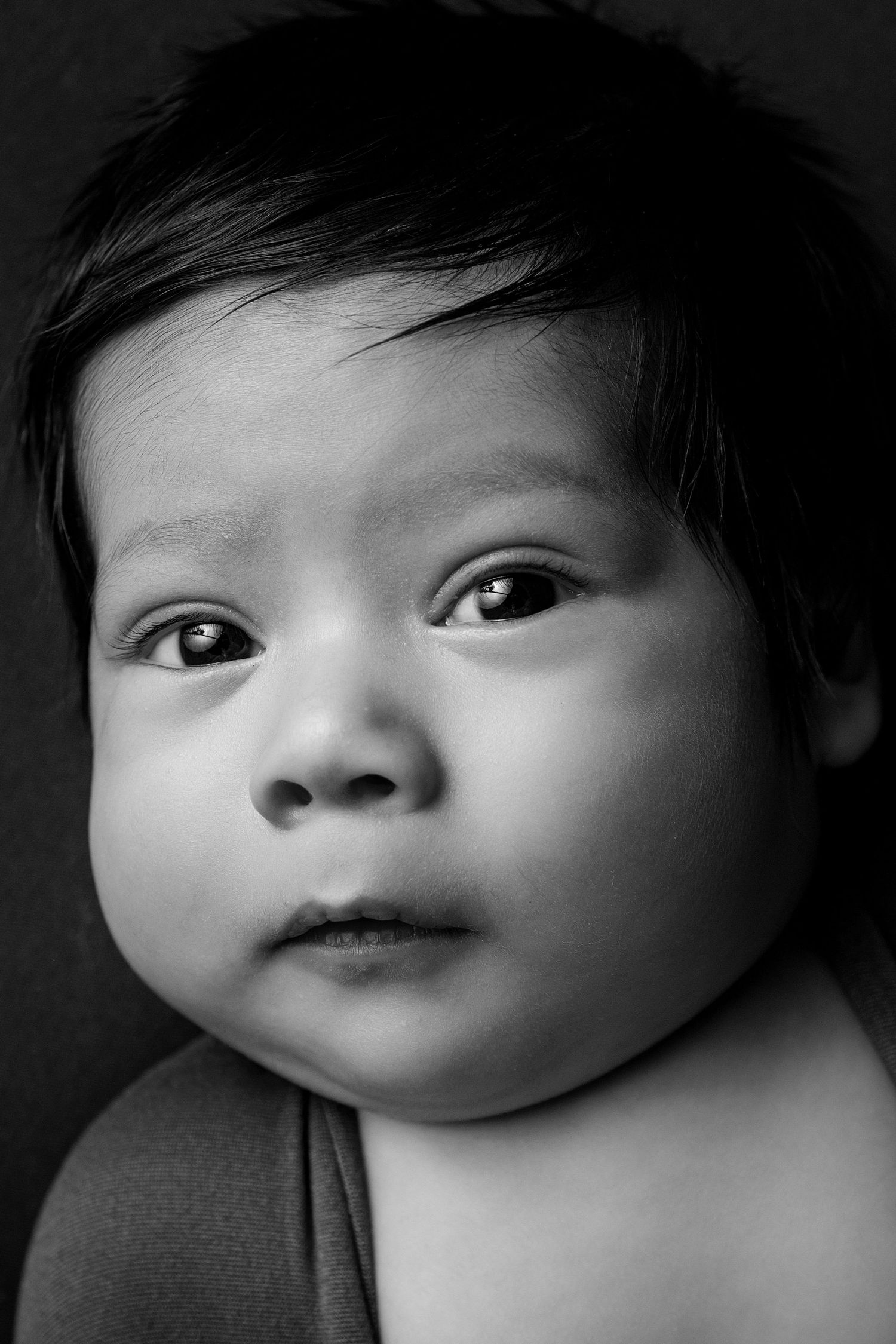Black and white portrait of an alert newborn baby with dark hair and wide eyes looking at the camera.