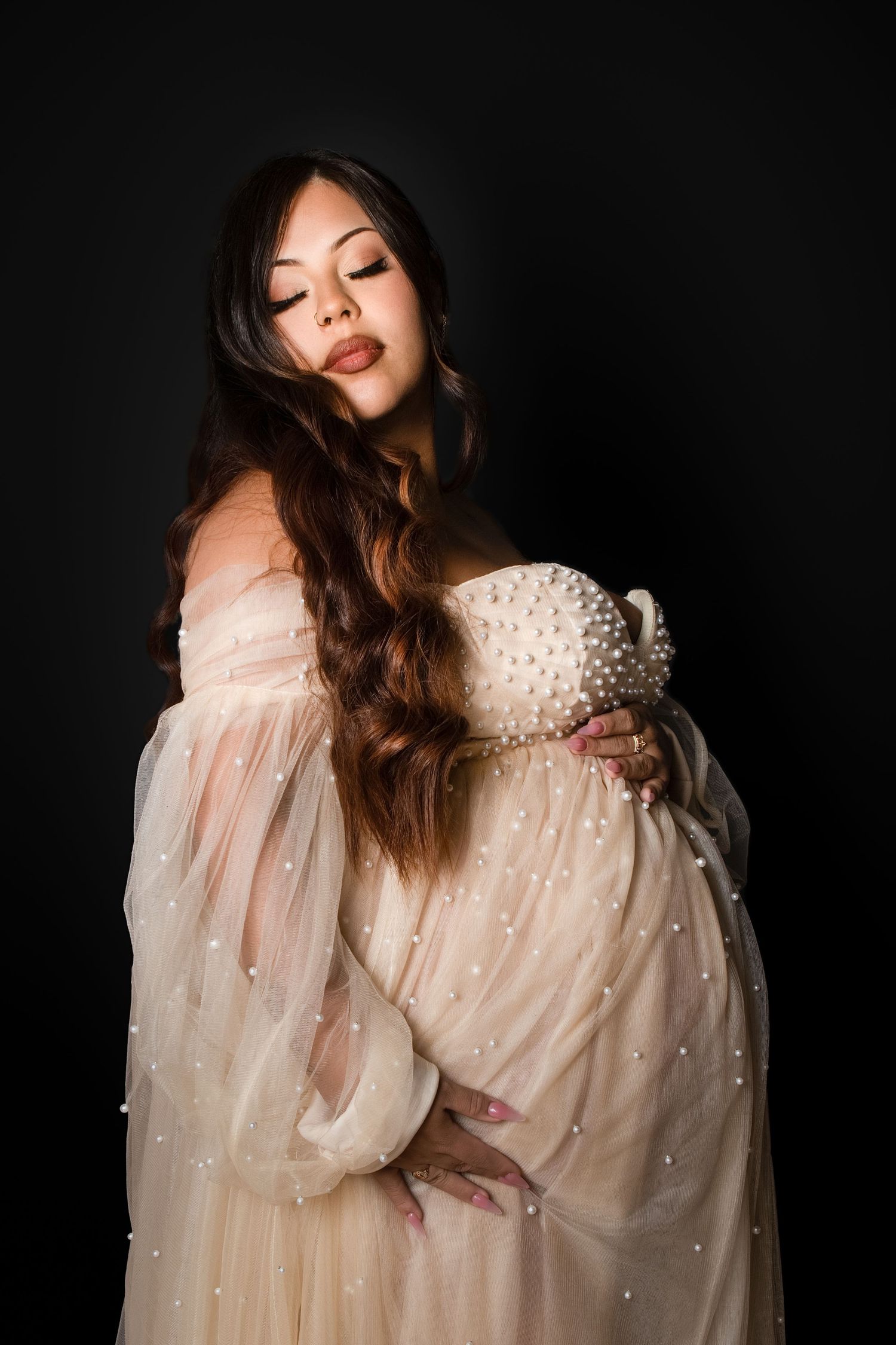 Pregnant woman in pearl-embellished sheer cream gown cradling baby bump against dark background.
