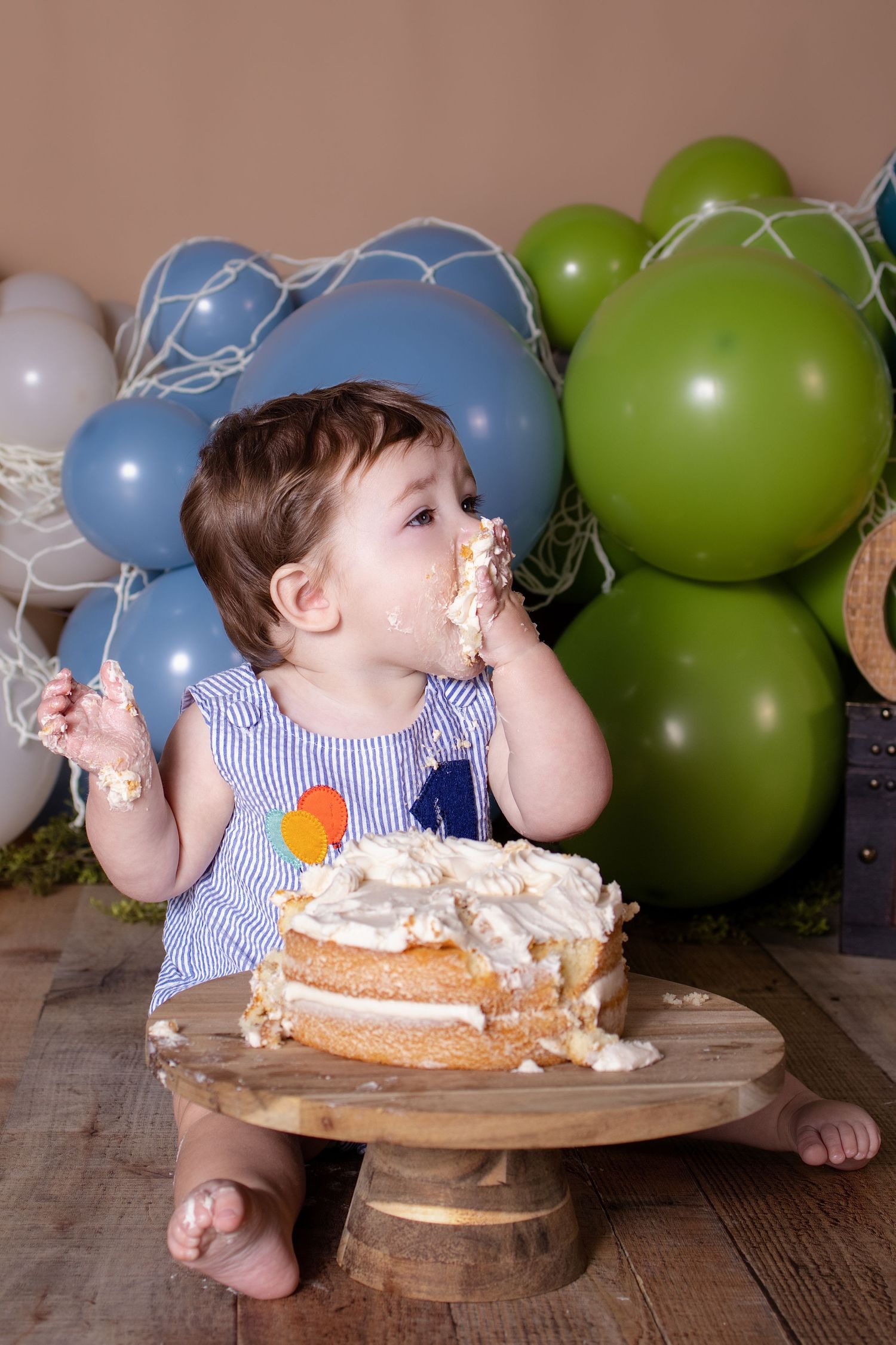 Baby in striped outfit eating first birthday smash cake with hands surrounded by blue and green balloon decor.